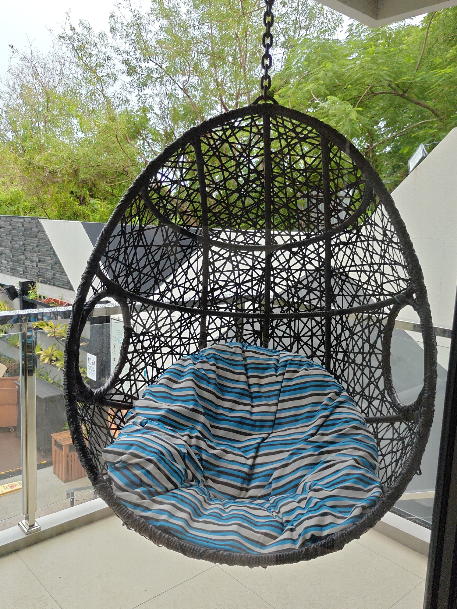 This image shows an outdoor hanging rattan chair with a blue, white, and black striped cushion, suspended by a black chain. The chair is situated on a balcony or patio, with a glass railing and a tiled floor visible in the foreground. In the background, lush green trees and a bright sky suggest a tropical setting. The architectural elements, including a white wall and a section of grey stonework, imply a modern building. The overall ambiance is one of relaxation and comfort, typical of leisure spaces in Pattaya City, Thailand, known for its resorts and outdoor living areas.