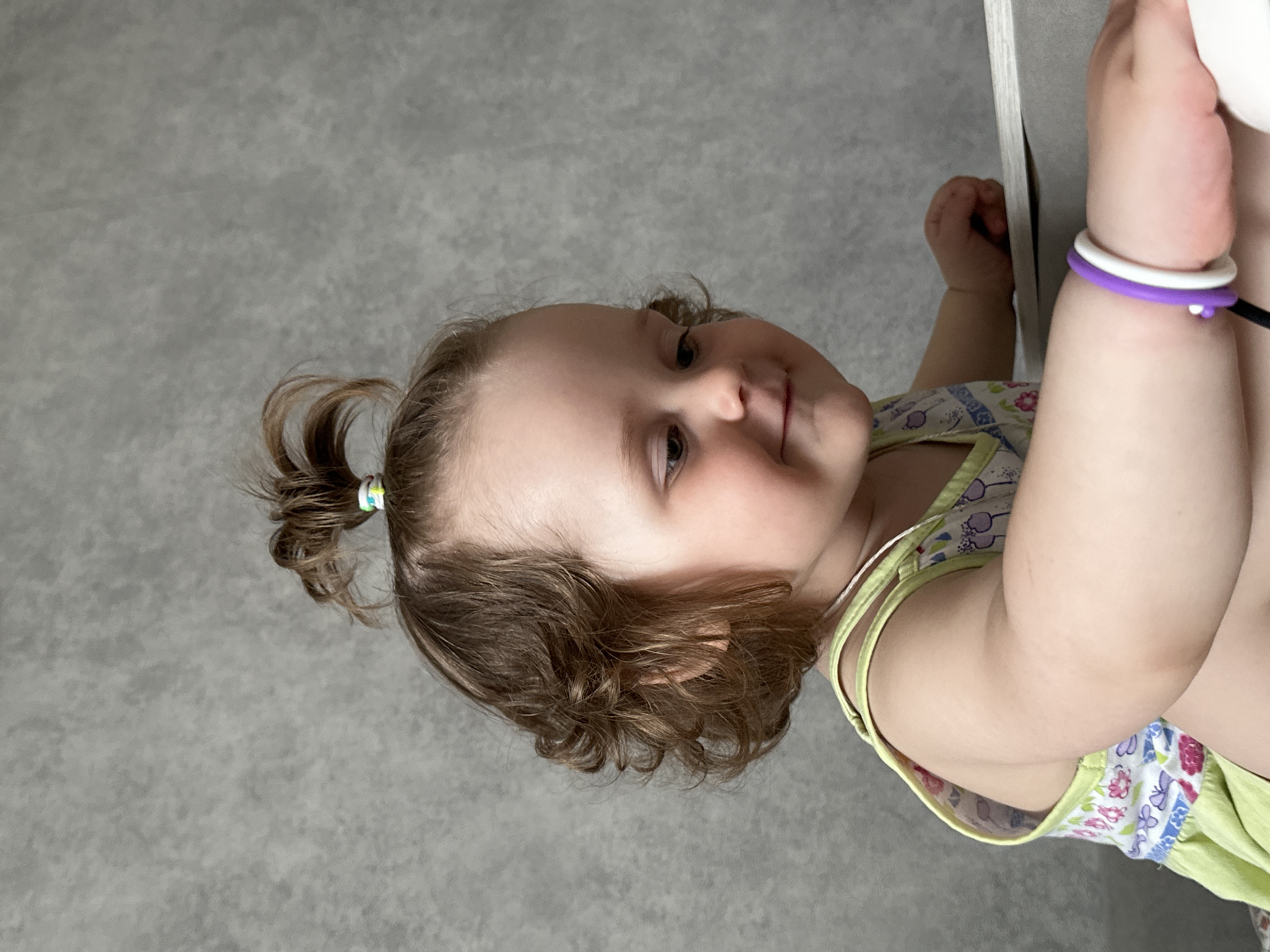 A toddler with brown curly hair, partially styled in a small ponytail, is pictured in a close-up shot. The child is wearing a light green and floral patterned tank top and has two bracelets on their left wrist, one white and one purple. The toddler is holding onto a white surface or edge, possibly a piece of furniture. The background is a textured gray wall. The location is stated to be Chaikovsky, Russia.