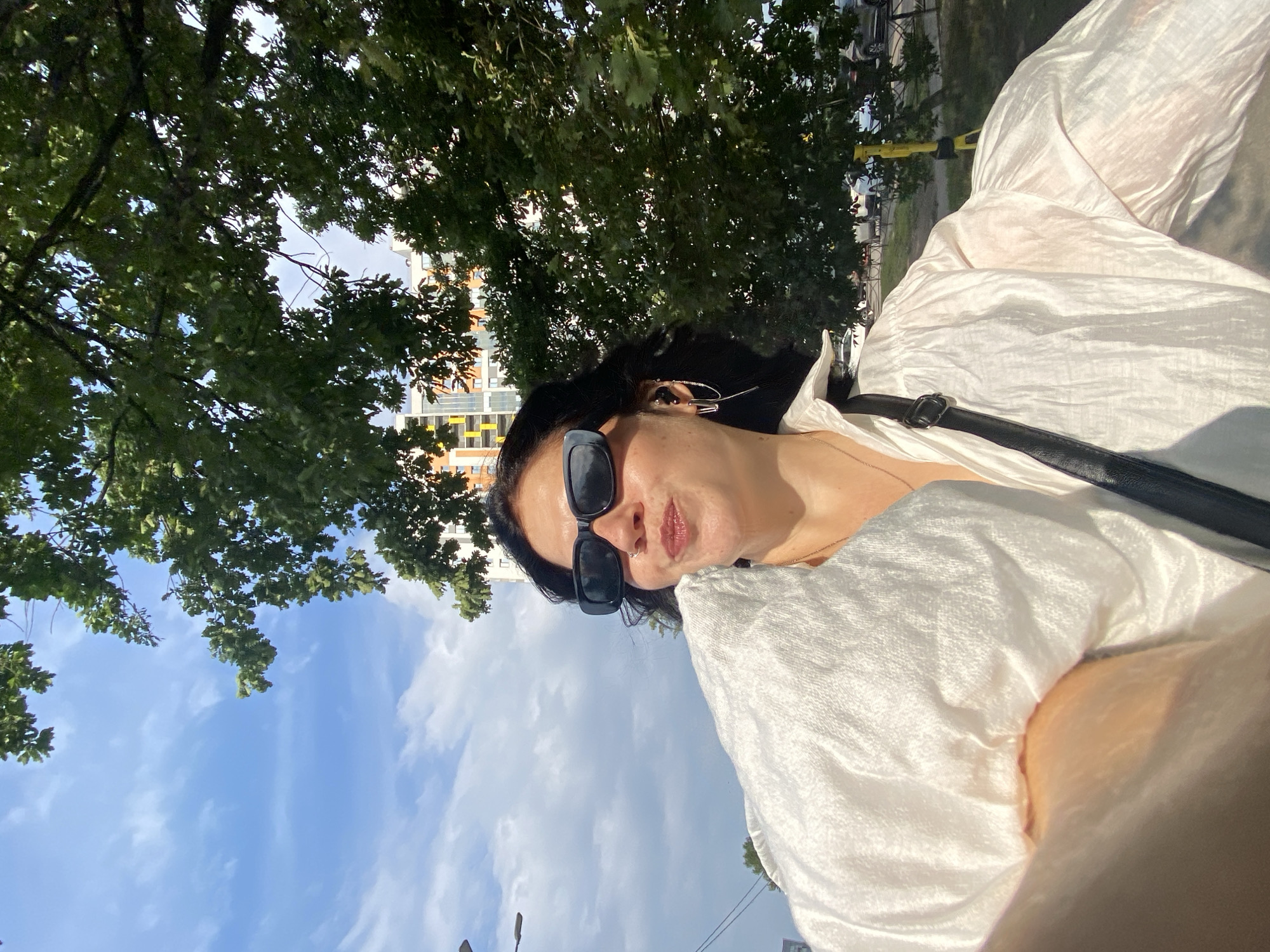 A woman with dark, wavy hair and black sunglasses is pictured from the chest up, taking a selfie outdoors. She is wearing a white, oversized, textured shirt and a black crossbody bag. She has on large hoop earrings and earbuds. In the background, a modern, colorful building is visible behind lush green trees. The sky is blue with scattered white clouds. The scene appears to be in an urban park or street area in Russia.