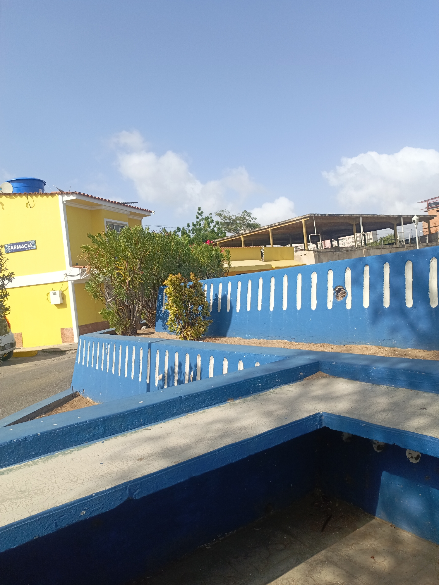 The image depicts a street scene in Juan Griego, Venezuela. A bright yellow building with a sign reading "Farmacia" in blue lettering is visible on the left, topped with a blue water tank. Adjacent to the building, a vibrant blue wall with vertical white stripes curves along the sidewalk. Behind the wall, there are bushes and a partially visible structure with a roof. The foreground features a multi-tiered concrete structure painted blue and white, with shadows cast inside it. The sky above is blue with scattered white clouds.