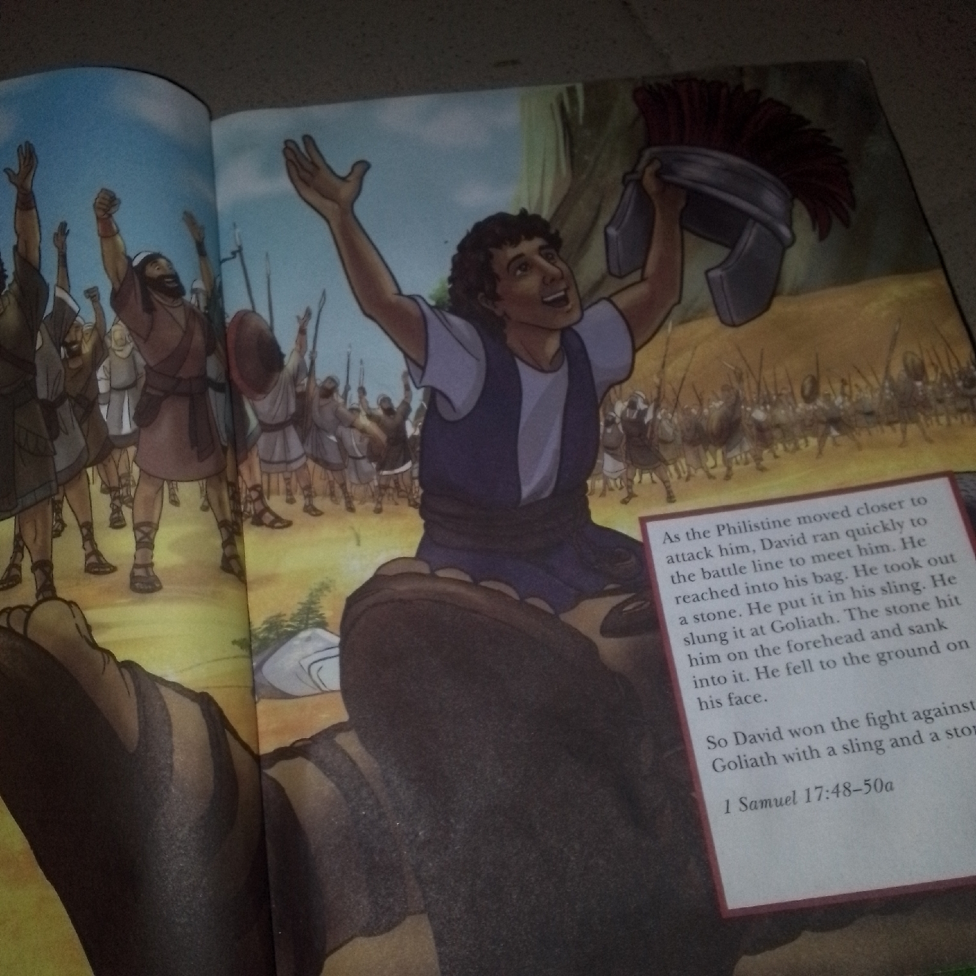 This media file shows an open children's book with an illustration depicting a scene from the biblical story of David and Goliath. The illustration is in a cartoon style and features two distinct parts. On the left, a crowd of people, presumably soldiers, are raising their arms in the air, suggesting a moment of triumph or anticipation. They are dressed in what appears to be ancient attire, with some carrying shields and spears. The background of this section is a bright blue sky.
The right side of the illustration focuses on a young man, identified as David by the text, who is holding up a large helmet with a red plume. He has a joyful expression on his face, with his arms outstretched. Behind him, another crowd of people, likely the Philistines, are depicted, appearing to be in battle formation with spears. The setting seems to be an outdoor battlefield, with some trees and a rocky landscape visible in the background.
A text box with a red border is placed in the lower right corner of the image, containing a narrative about the events depicted. The text describes David's actions, his quick approach to the battle line, taking a stone, putting it in his sling, and hitting Goliath. It also mentions Goliath falling to the ground and David winning the fight. The text concludes with a Bible verse reference: "1 Samuel 17:48-50a". The lighting suggests it is daytime. The image is a close-up of the book, with parts of the surrounding environment, like the page edges and possibly a floor or table, visible at the edges.