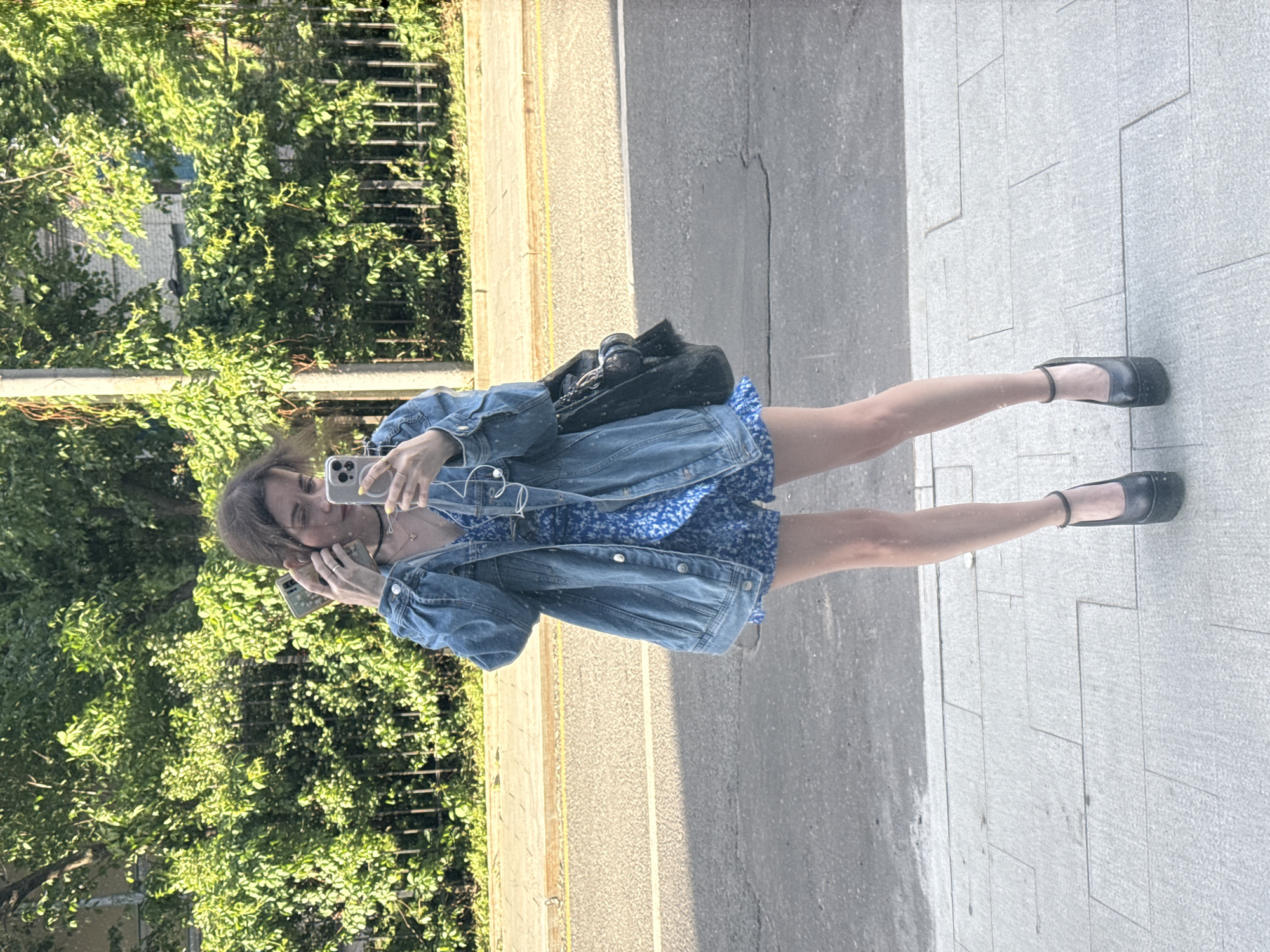 A young woman stands on a sidewalk in what appears to be Moscow, Russia, based on the Cyrillic text visible in reflections. She is taking a selfie with her smartphone. The woman is wearing a blue floral dress and a denim jacket. She has a black bag slung over her shoulder and is wearing black flat shoes. The environment includes trees, a street, and a paved area.