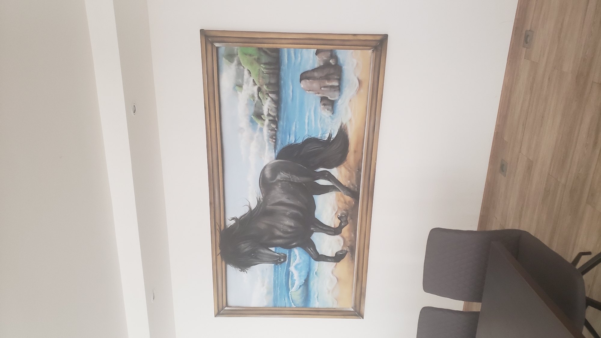 The image displays a framed painting of a black horse running on a beach with the ocean in the background. The painting is hung on a white wall, above two dark gray chairs and a dark brown table. The floor is made of light brown wooden planks. The painting is rectangular and the frame is a light brown color. The painting itself depicts a dark horse with a flowing mane and tail, its front hooves raised as it runs along the sandy shore. The ocean is a vibrant blue with white waves crashing onto the beach. In the distance, there are green hills and rocky outcrops. The scene is brightly lit, suggesting an indoor setting with good lighting. There are no people visible in the image. The image quality is good, allowing for clear observation of the details in the painting and the surrounding environment. There is no information available to determine the specific location in the city of Prešov, Slovakia.