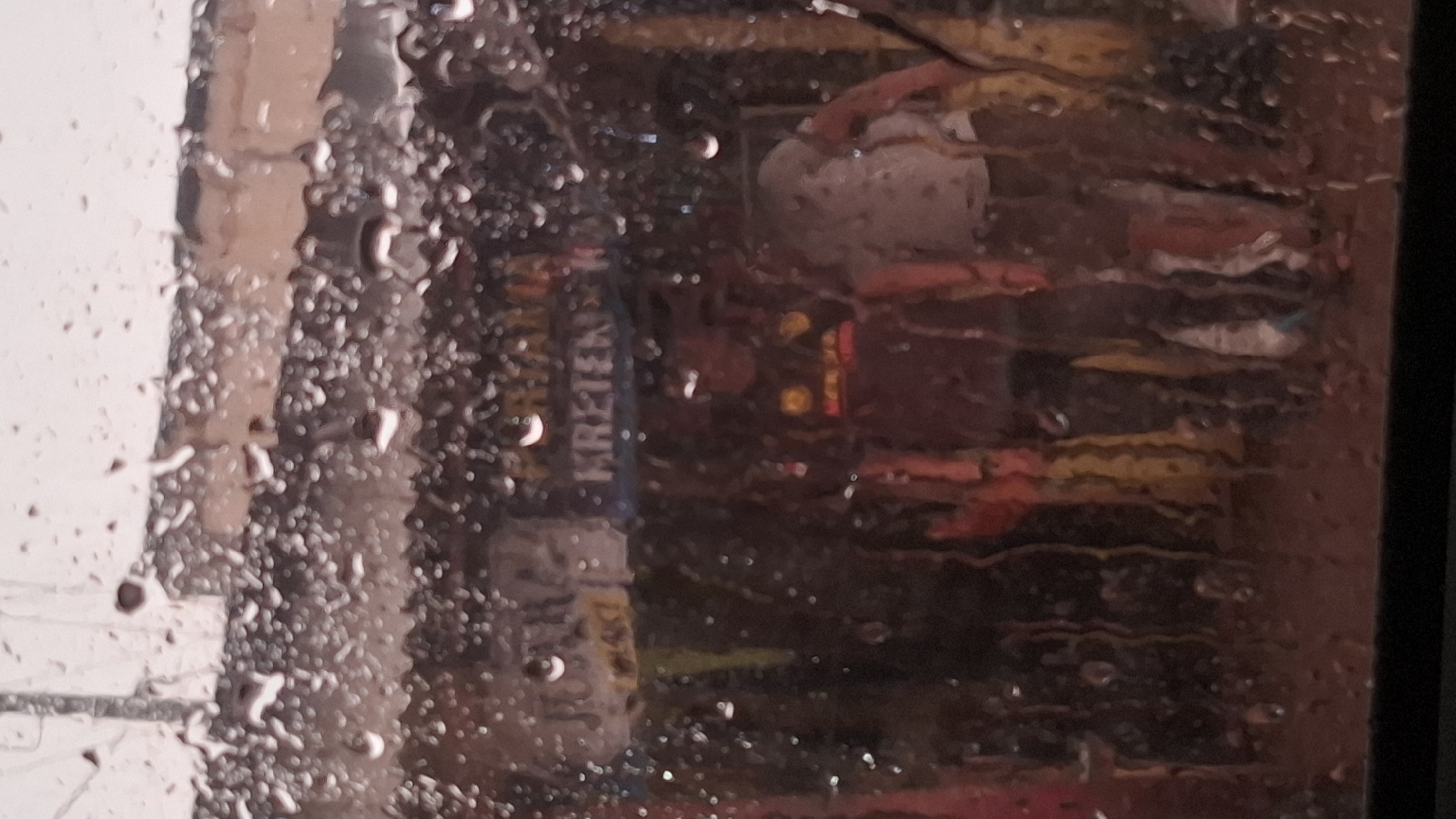 The image depicts a scene through a rain-streaked window, suggesting it was taken from inside a vehicle or building. The foreground is dominated by numerous water droplets distorting the view of the background. In the background, there appear to be people standing in what might be an outdoor market or street setting. Two individuals are more clearly visible, one wearing a dark shirt with a yellow emblem and the other in a lighter, possibly patterned, shirt. Signs are visible behind them, with text that is partially obscured by the water and the blurriness of the image. One sign appears to have the word "KRETEK" on it, which is a type of Indonesian clove cigarette. Another sign has illegible writing. The overall impression is one of a slightly chaotic, possibly commercial, environment. The mention of Cilaku, Indonesia, implies this scene is located in that specific region.