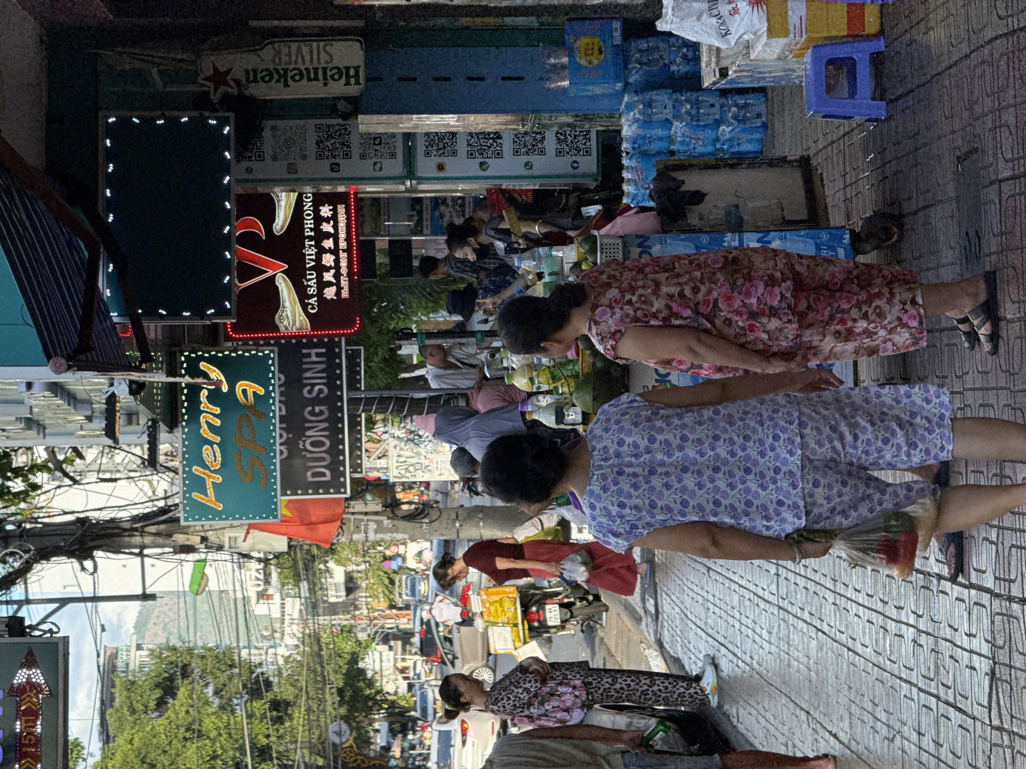 This image depicts a street scene in Nha Trang, Vietnam, likely a commercial area with shops and signage lining the sidewalk. In the foreground, two women are walking away from the camera on a patterned tiled sidewalk. The woman on the left wears a leopard-print outfit and carries a large patterned bag. The woman on the right wears a floral print outfit and carries a clear plastic bag containing what appear to be fruits and vegetables. Several other people are visible in the mid-ground, some appearing to be shoppers or vendors.
Behind the people, a variety of signs are displayed. One prominent sign is blue with yellow lettering that reads "Henry SPA". Below this, another sign is dark with white lettering that says "DUONG SINH". To the right, a neon sign features a red "V" symbol above text in Chinese and Vietnamese, including "CÁ SẤU VIỆT PHONG" and "越风鳄鱼皮料" (Viet Phong Crocodile Leather Goods). Further to the right, a tall, vertical Heineken Silver sign is visible.
The environment includes a street with light traffic, including cars and motorcycles, suggesting an urban setting. There are overhead power lines and a mix of buildings visible in the background. Stalls or shops are set up along the sidewalk, with goods like bottled water and crates of products displayed. A small blue plastic stool is placed on the sidewalk near some of these stalls. The overall impression is a bustling street market or commercial street.