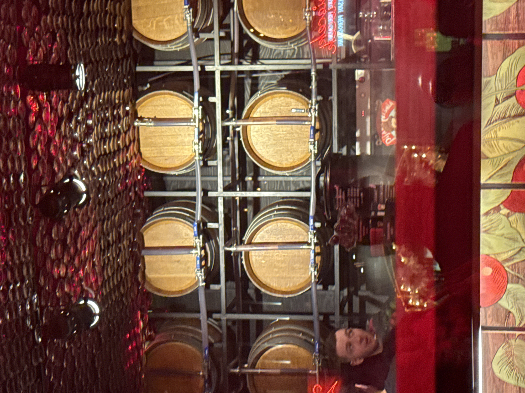 In an interior scene, rows of wooden barrels are stacked on metal shelving units. The barrels are light brown with dark metal bands. Above the barrels, the ceiling is covered with a decorative, textured material that appears to be lit from behind with red lights, creating a shimmering effect. In the foreground, a young man with short brown hair and a beard is partially visible on the left side of the frame, looking to the right. He is wearing a dark shirt. A red reflective surface is in front of the man, obscuring the lower portion of the barrels and creating reflections. In the background, behind the barrels, shelves with bottles and other items are visible, along with signage.