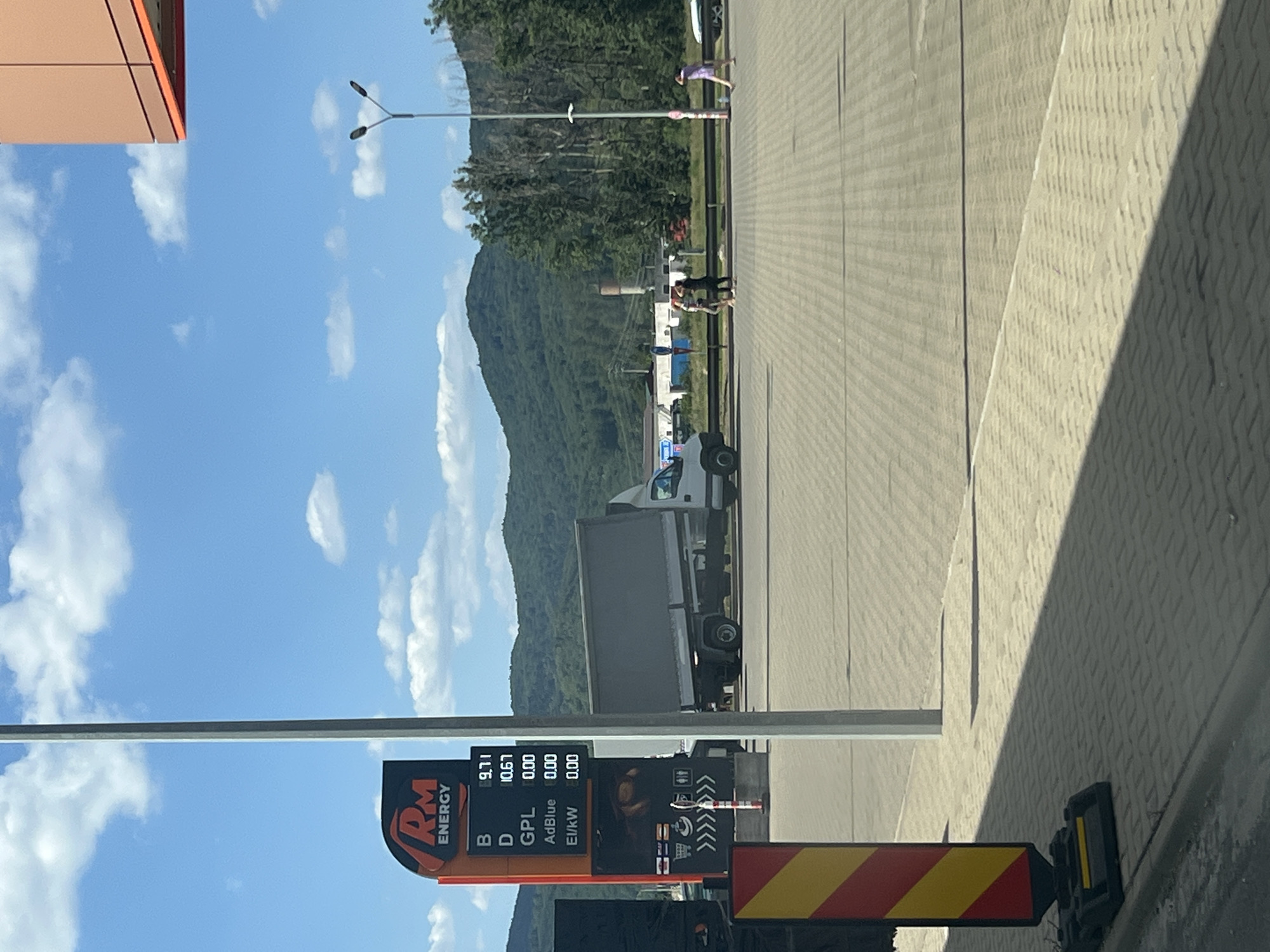 The image shows a gas station sign at RM Energy in Păltinoasa, Romania, on a sunny day with a blue sky and scattered clouds. The sign displays fuel prices: "B 9.71", "D 10.67", "GPL 0.00", "AdBlue 0.00", and "El/kW 0.00". A gray truck is parked to the right of the sign. In the background, there are green hills and trees, and a tall industrial smokestack is visible. Several people are standing and walking in the mid-ground, and a lamppost is present. The foreground shows a paved area with shadows cast by a pole and a yellow and red striped marker.