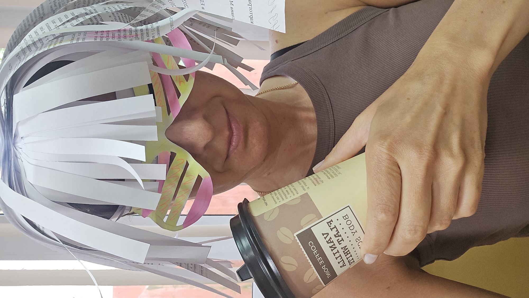 A woman in a brown tank top holds a coffee cup with text "BODY 20" printed on it. She is wearing a headpiece made of white and colored paper strips. The location appears to be indoors, with a window visible in the background. The city and specific location are not discernible.