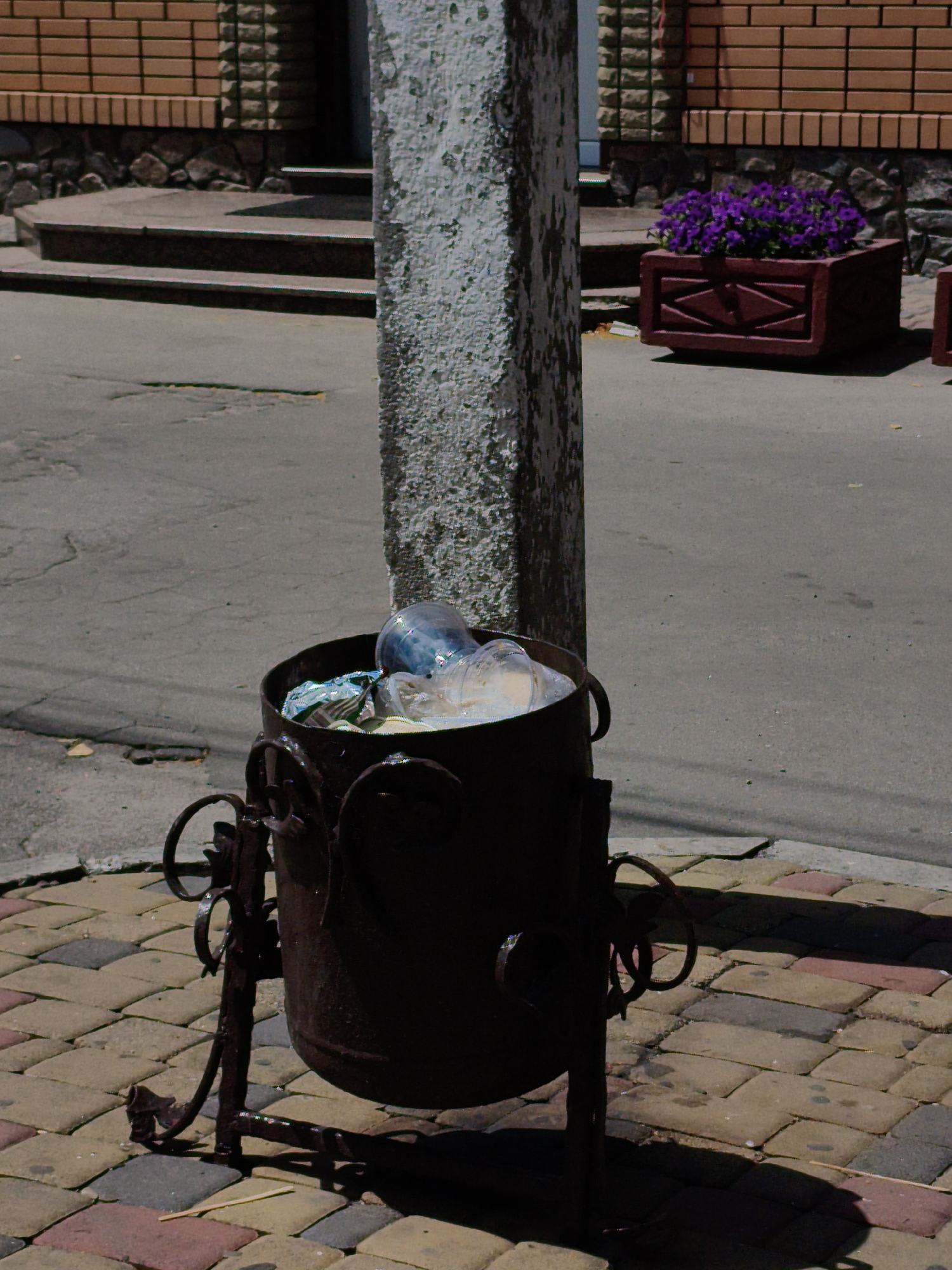 The image depicts an outdoor scene on a sunny day. In the foreground, a dark, weathered metal trash can with ornate wrought-iron legs stands on a multi-colored paved walkway. The trash can is partially filled with plastic bottles and bags. Behind the trash can, a rough, textured concrete pillar is visible. The walkway transitions to a grey asphalt surface, which appears slightly cracked. In the background, a building with a brick facade and stone foundation is present, with a set of stairs leading up to an entrance. To the right of the stairs, a dark red planter box filled with vibrant purple flowers adds a splash of color to the scene. This image does not provide any specific indicators of the location being in Velyka Bahachka, Ukraine. There are no people or animals visible in the frame.