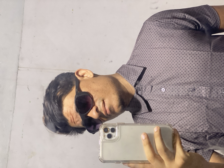 A man with dark hair wears sunglasses and a patterned gray shirt. He angles his head to the side, holding a phone with a clear case in his hand. The background appears to be a plain white wall. The image seems to be a self-portrait or a photo taken with a timer.