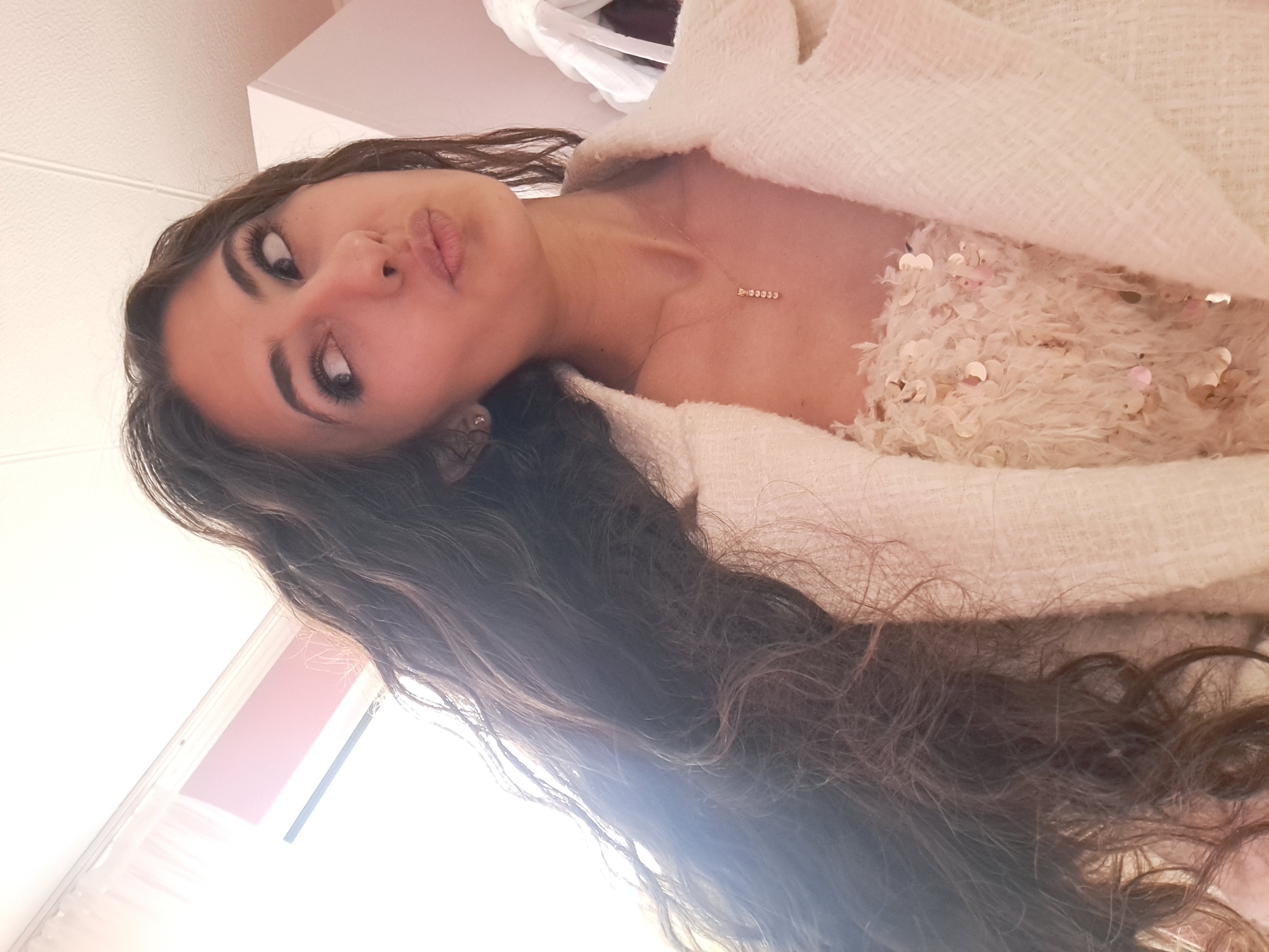 A young woman with long, dark, curly hair is looking upwards and to the right, with her lips pursed. She is wearing a white tweed jacket over a light-colored, sequined top and a delicate necklace. The lighting suggests she is indoors, possibly near a window. There is no visible indication of the location being in the city of Negovan, Bulgaria.