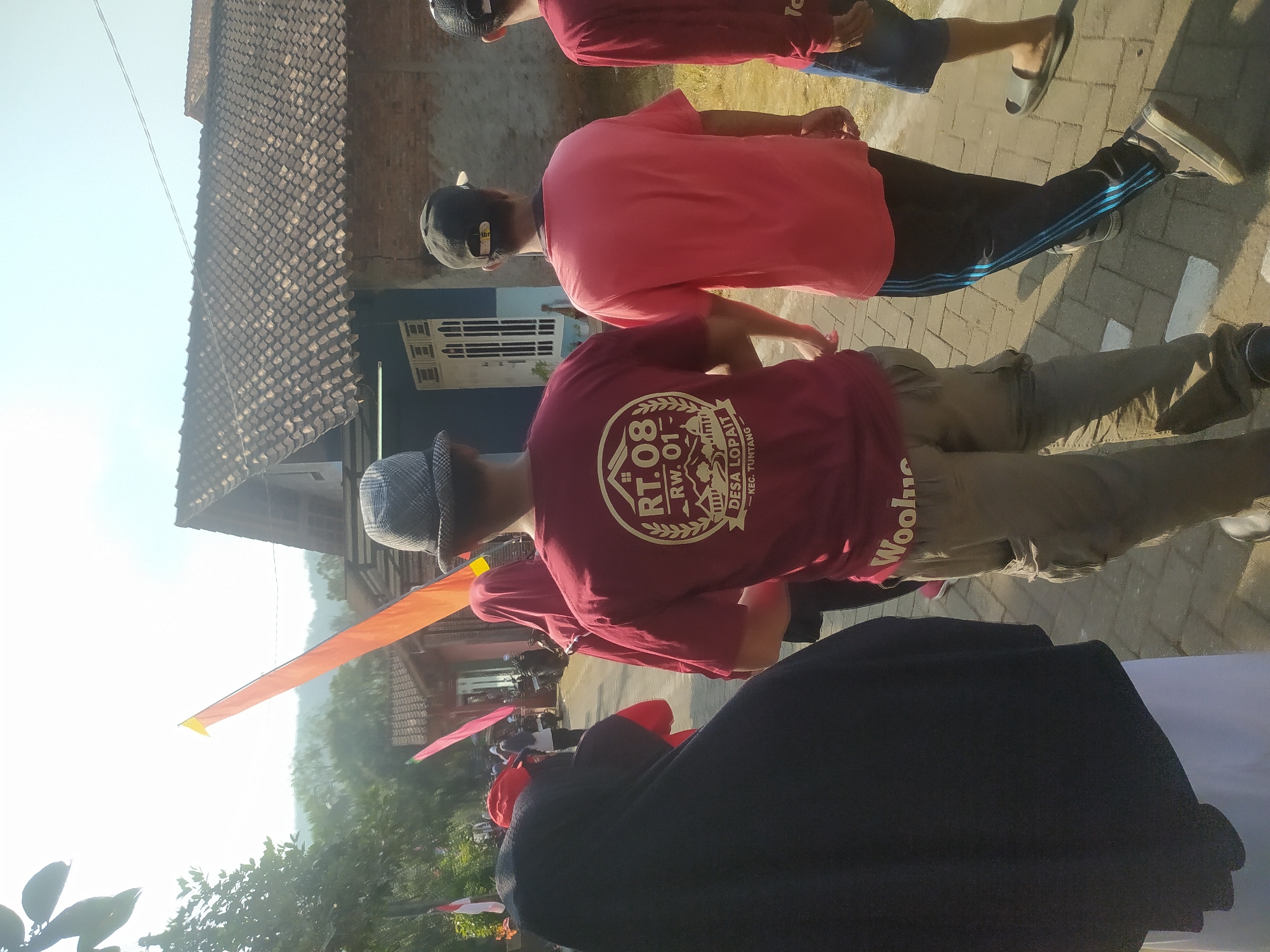 A group of people are walking down a brick pathway in the village of Lopait, Indonesia. The group includes individuals wearing a black hijab, a maroon t-shirt with a circular logo that reads "RT.08 RW.01 DESA LOPAIT KEC. TUNTANG", a pink t-shirt, and a red long-sleeved shirt. They are walking away from the camera, towards houses with tiled roofs. In the background, orange and pink flags are visible. The environment appears to be rural and sunny, with trees and hills in the distance.