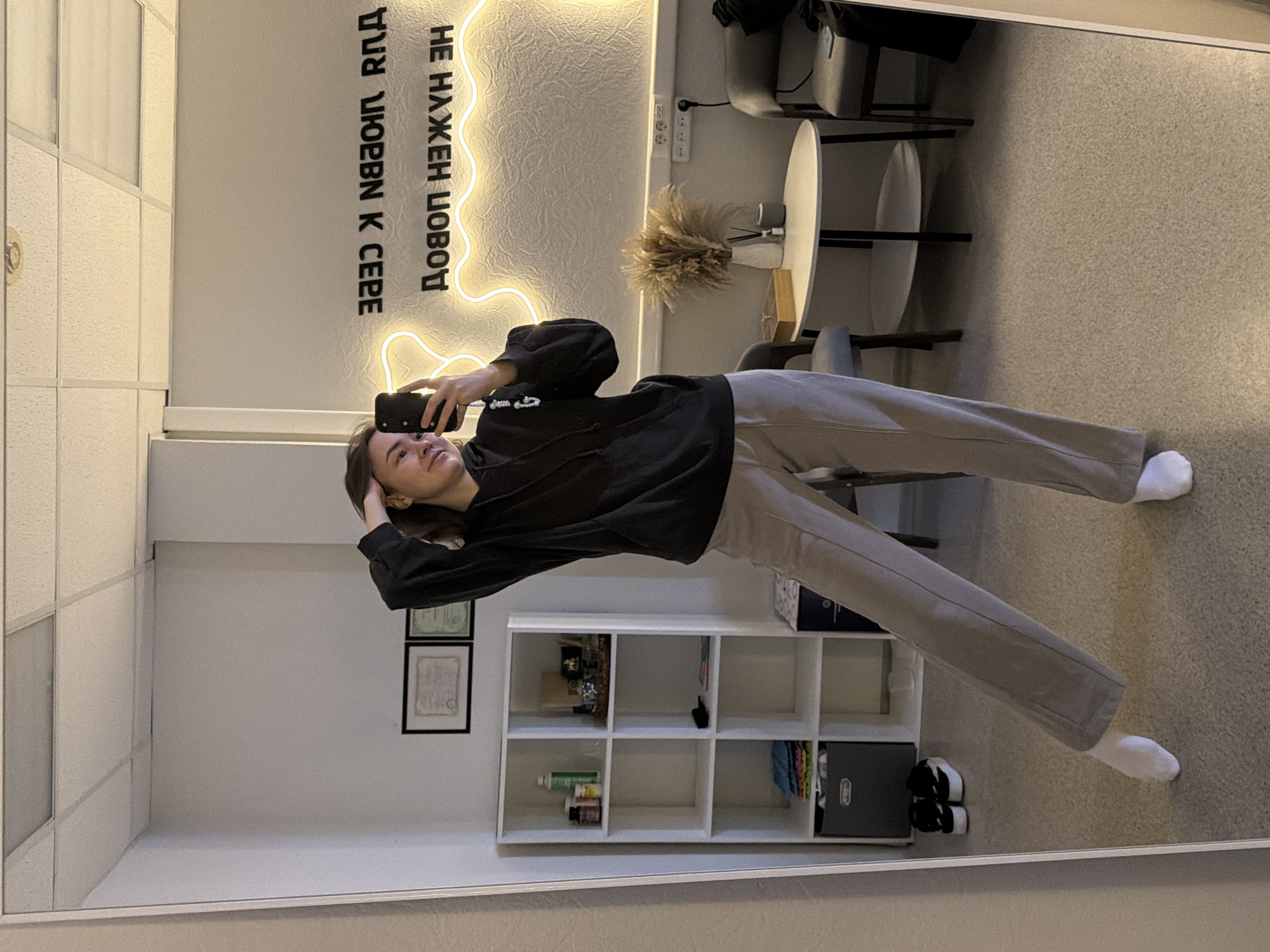 The image shows a reflection in a mirror. A woman is holding a mobile phone up, presumably taking a photograph of herself. She is wearing a black hoodie and light-colored sweatpants with white socks. She is standing in front of a wall on which is a neon sign. Also visible in the room are chairs, a table, shelves containing various objects, and two framed documents on the wall. The image was likely taken in Chaikovsky, Russia, based on the writing on the neon sign.