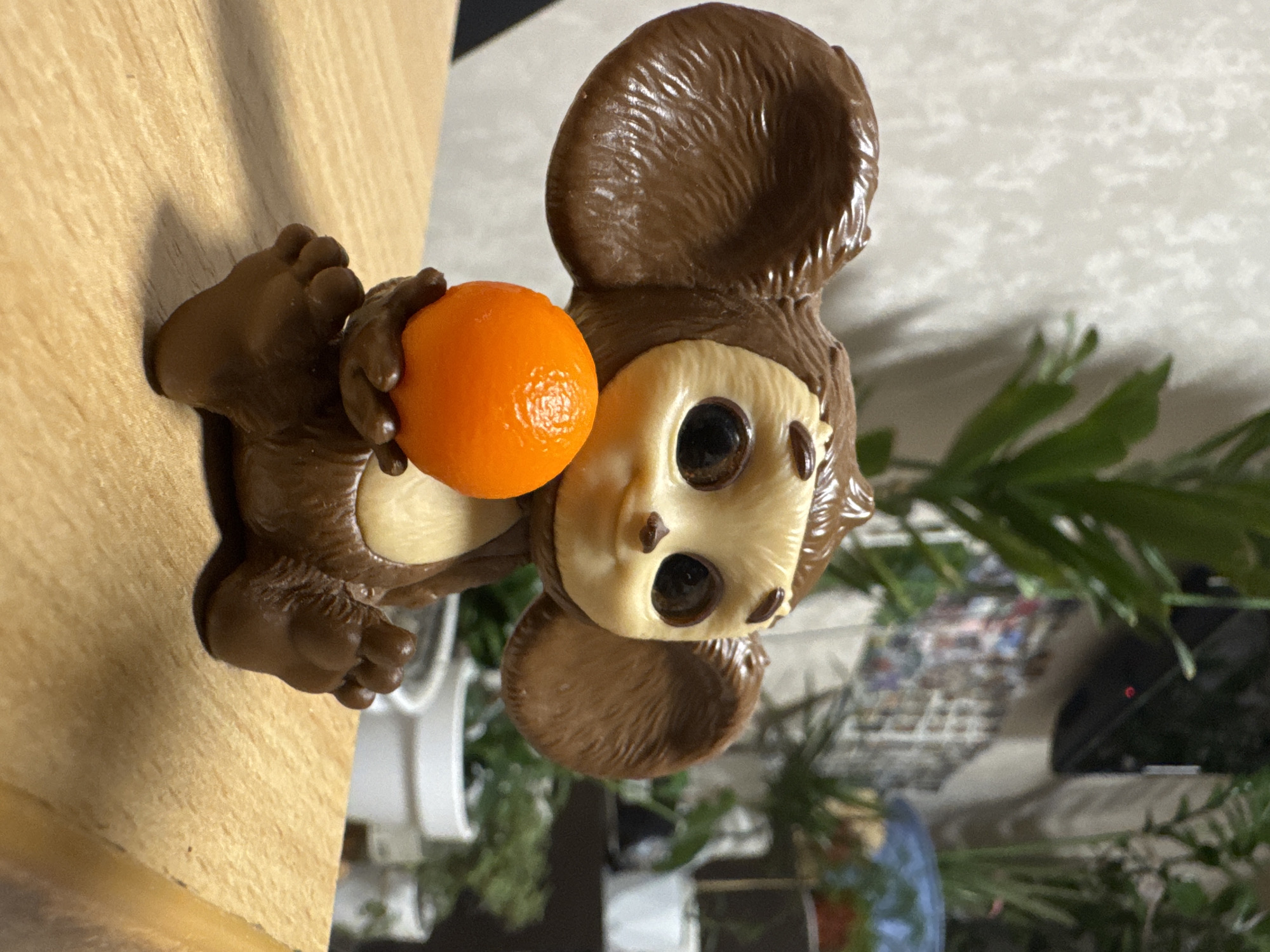 The image features a Cheburashka figurine, a character from Russian animation, placed indoors in Izhevsk, Russia. The figurine is predominantly dark brown with large, rounded ears and small, bare feet with defined toes. Its face is a lighter, beige color with large, dark, round eyes and small brown details. The figurine holds a small, vibrant orange fruit, appearing to be a tangerine or clementine, in its hands.
The setting is an indoor environment, with the figurine resting on a light-colored wooden surface, possibly a table or shelf. The background is softly blurred, showing green foliage from a houseplant and other indistinct objects, including a white container and a dark item with a small red light. The scene is static, depicting the figurine in a posed position. The lighting indicates it is likely daytime indoors. There is no visible text.