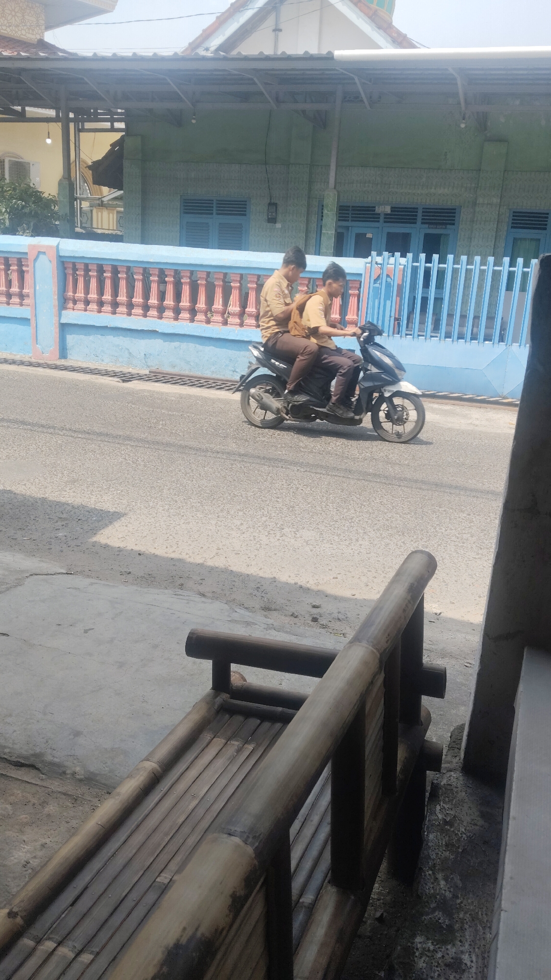 Two male students in tan uniforms ride a motorcycle on a paved road in Bandar Lampung, Indonesia. The student in front is driving, and the student in the back is wearing a backpack. They are passing a blue and pink fence. In the background, there is a light green building with blue windows and a pitched roof. In the foreground, a bamboo bench is visible. The scene is outdoors during the day, with bright sunlight casting shadows.