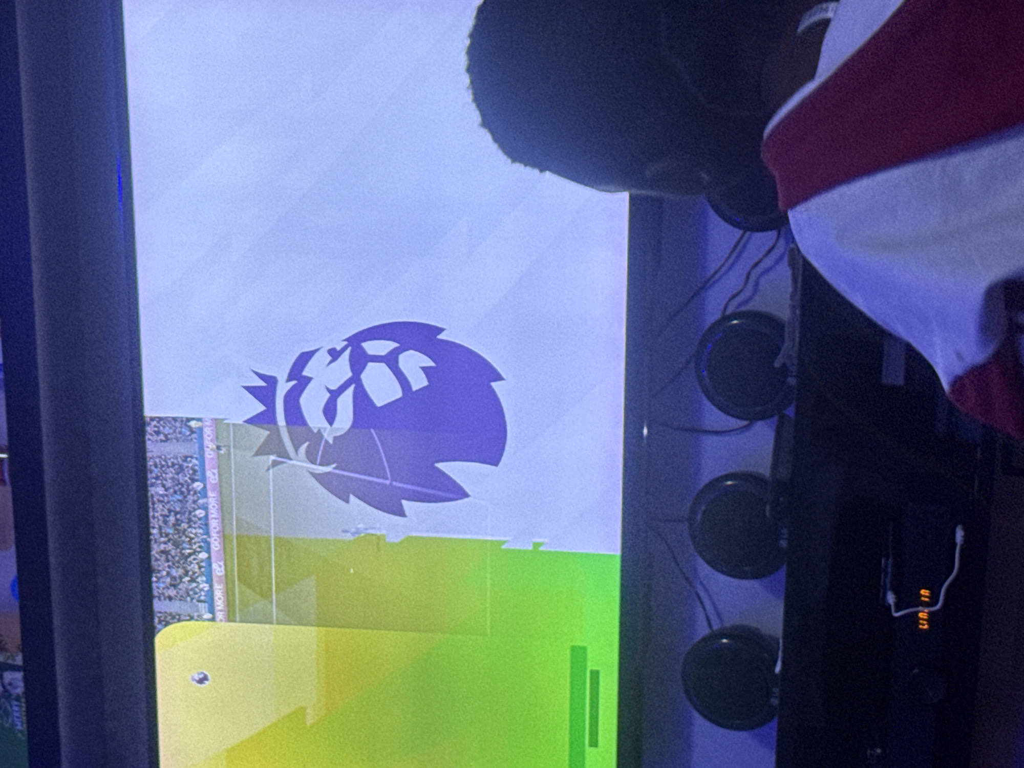 A television screen displays the Premier League logo over a green and yellow graphic, possibly from a video game or broadcast. To the right, a person's head and shoulder, partially obscured, are visible, wearing a white and maroon garment. Behind the television, wires and round objects, likely speakers or microphones, are arranged vertically. There is no information available about Jos, Nigeria.