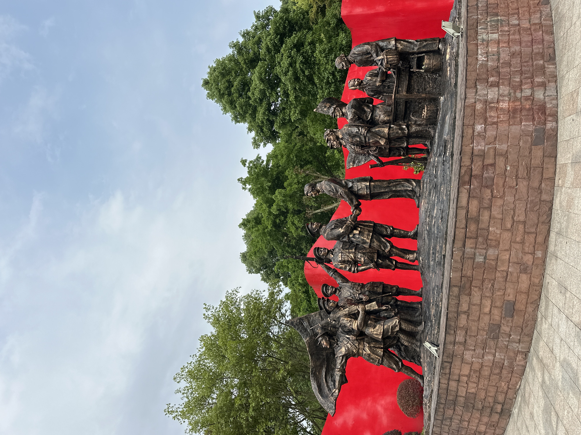 The image shows a bronze sculpture against a bright red backdrop. The sculpture depicts a group of people, possibly soldiers, in various action poses. Trees and a cloudy sky are visible in the background. The base of the sculpture appears to be made of brick. The location is the city of Liangjiang, China.