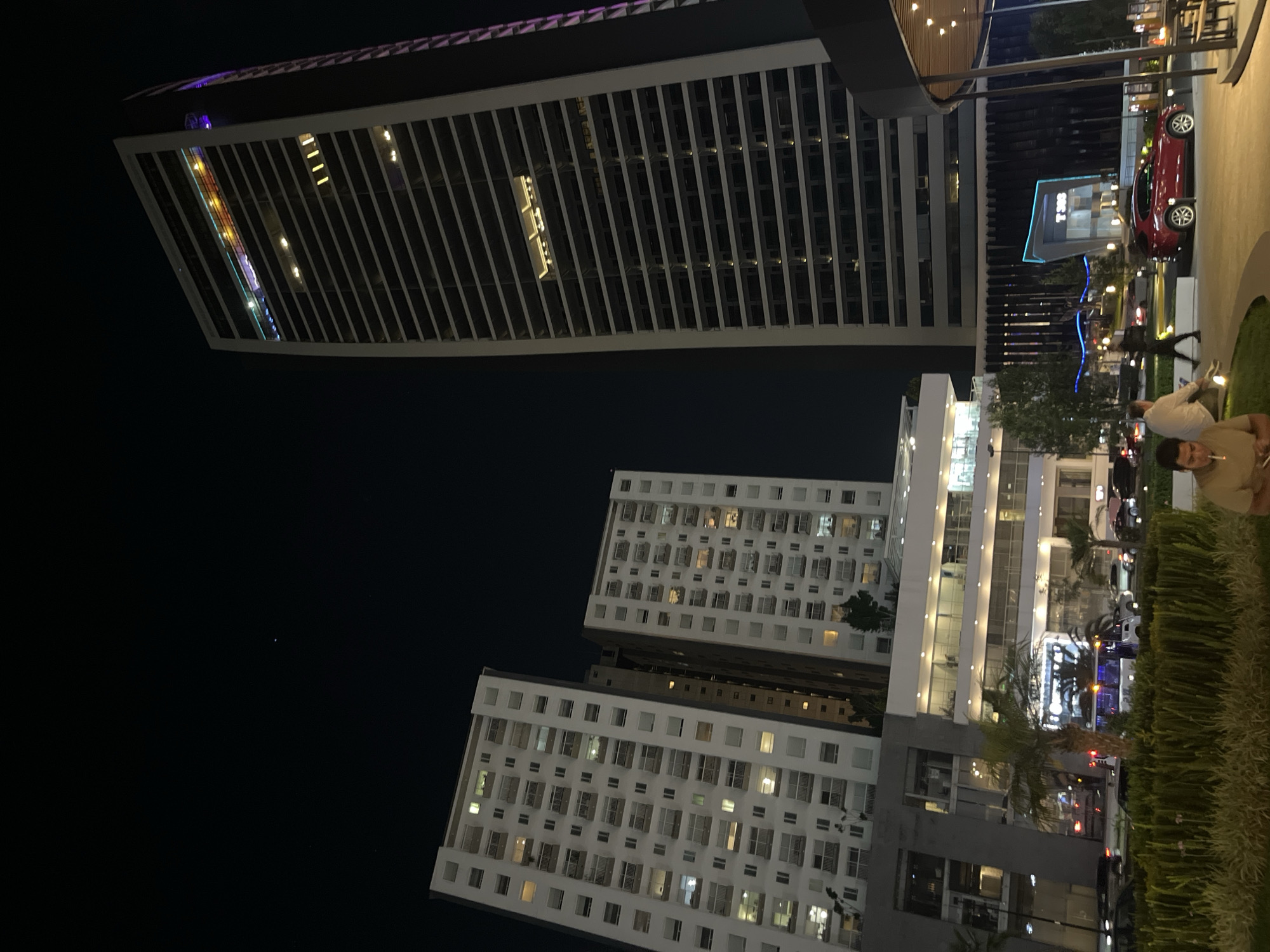 The media depicts an outdoor urban night scene in San Bernardino Tlaxcalancingo, Mexico, showcasing a mix of modern and more traditional architecture.
A very tall, modern skyscraper with a distinctive angled top and prominent vertical strip lighting dominates the left side of the frame (when viewed upright). The building's windows reflect interior lights and possibly other light sources. Faint, illuminated text, possibly "BBVA", is visible on the side of this skyscraper but is not clearly legible.
To the right of the skyscraper are two multi-story buildings, likely residential or older office blocks, characterized by numerous windows, many of which are illuminated from within, and some featuring shutters.
In the mid-ground, a modern structure with bright blue accent lighting stands out, possibly a commercial entrance or retail space. A red car is parked near this structure, and other vehicles are visible on a street in the background.
Several people are present in the scene, suggesting an active area. Some individuals are seated on a low wall or bench in the foreground, while others are standing or walking. One person in a light-colored shirt is partially visible in the bottom right foreground, looking towards the viewer.
The time of day is clearly nighttime, with a dark sky illuminated primarily by the artificial lights of the buildings, streetlights, and some decorative string lights. Hedges and some small trees contribute to the landscaping of the outdoor space where people are gathered. No other notable text is clearly visible.