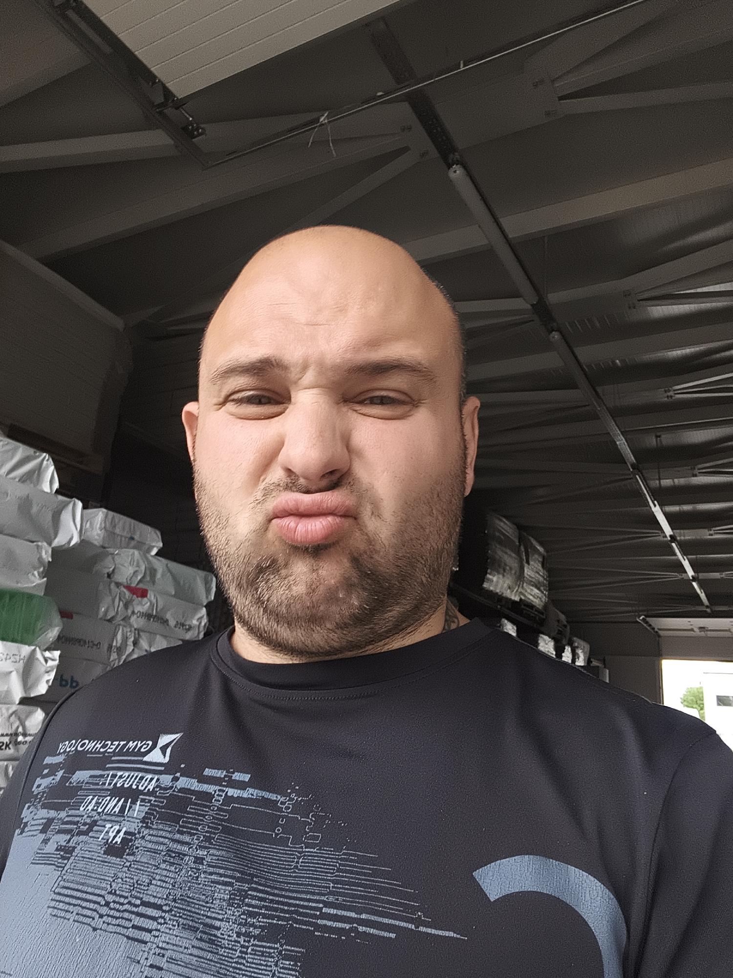This is a medium shot, selfie-style photograph of a bald man with a beard, making a duck face. He is wearing a dark t-shirt with a graphic design on it, featuring what appears to be circuitry or abstract lines in shades of blue and grey. The text on the shirt is partially visible and appears to be reversed, possibly indicating it's a reflection or the shirt is inside out.
The man's expression is playful or humorous. His skin appears to have some sweat or shine, suggesting it might be warm or he has been active.
The background is out of focus but shows what looks like a warehouse or storage facility. Stacked white sacks are visible to the left, and the ceiling structure with beams and lighting fixtures is prominent above. The lighting suggests an indoor setting, possibly daytime given the ambient light. There are no clear indications of the time of day, weather, or a specific geographical location. The overall scene is utilitarian and industrial.