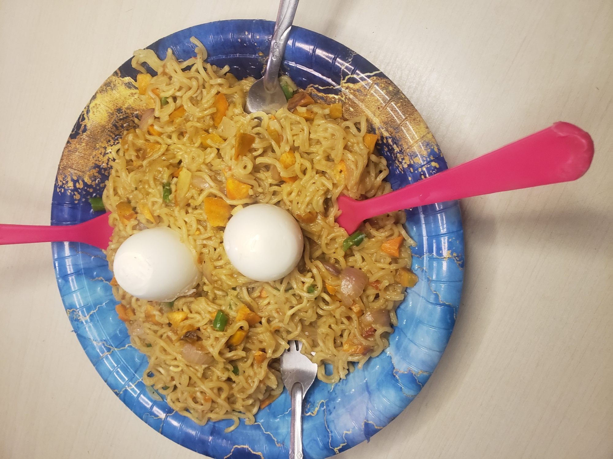 A close-up, overhead shot displays a blue, patterned disposable plate filled with instant noodles, topped with two boiled eggs. The noodles are mixed with diced carrots, green beans, and onions. Three plastic cutlery items are present: two pink spoons and one silver fork, two of which are partially submerged in the noodles. The plate rests on a light-colored, wood-grained surface, likely a table. The scene appears to be indoors.