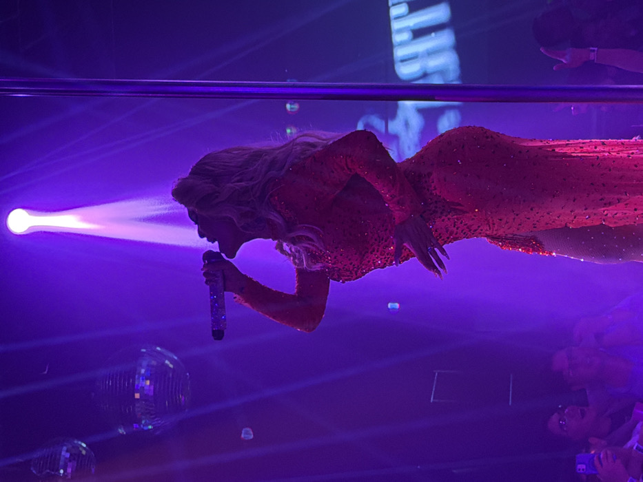 The image captures a dynamic scene from a live performance, likely a concert or drag show, set in an indoor venue in San Andrés Cholula, Mexico. The primary subject is a performer, seen from the chest upwards, dressed in a long, form-fitting, sparkling red gown adorned with sequins or glitter. They have long, light-colored hair and are intensely focused on a glittery microphone held close to their mouth, suggesting they are singing or speaking.
The stage is dramatically lit with a strong white spotlight beaming directly onto the performer from the left side of the frame. The ambient lighting is a rich purple, casting an atmospheric glow over the entire scene. On the left side of the image, two mirrored disco balls are visible, reflecting light and adding to the festive atmosphere. A horizontal bar, possibly part of the stage structure or lighting rig, spans the upper portion of the frame.
In the bottom right, several audience members are partially visible, looking upwards towards the stage, with some holding smartphones to record the event. Behind the performer, on a large screen or backdrop, faint, stylized white text is discernible but largely illegible. The overall impression is of an energetic nighttime performance, highlighting the performer's glamorous attire and the vibrant stage lighting.
