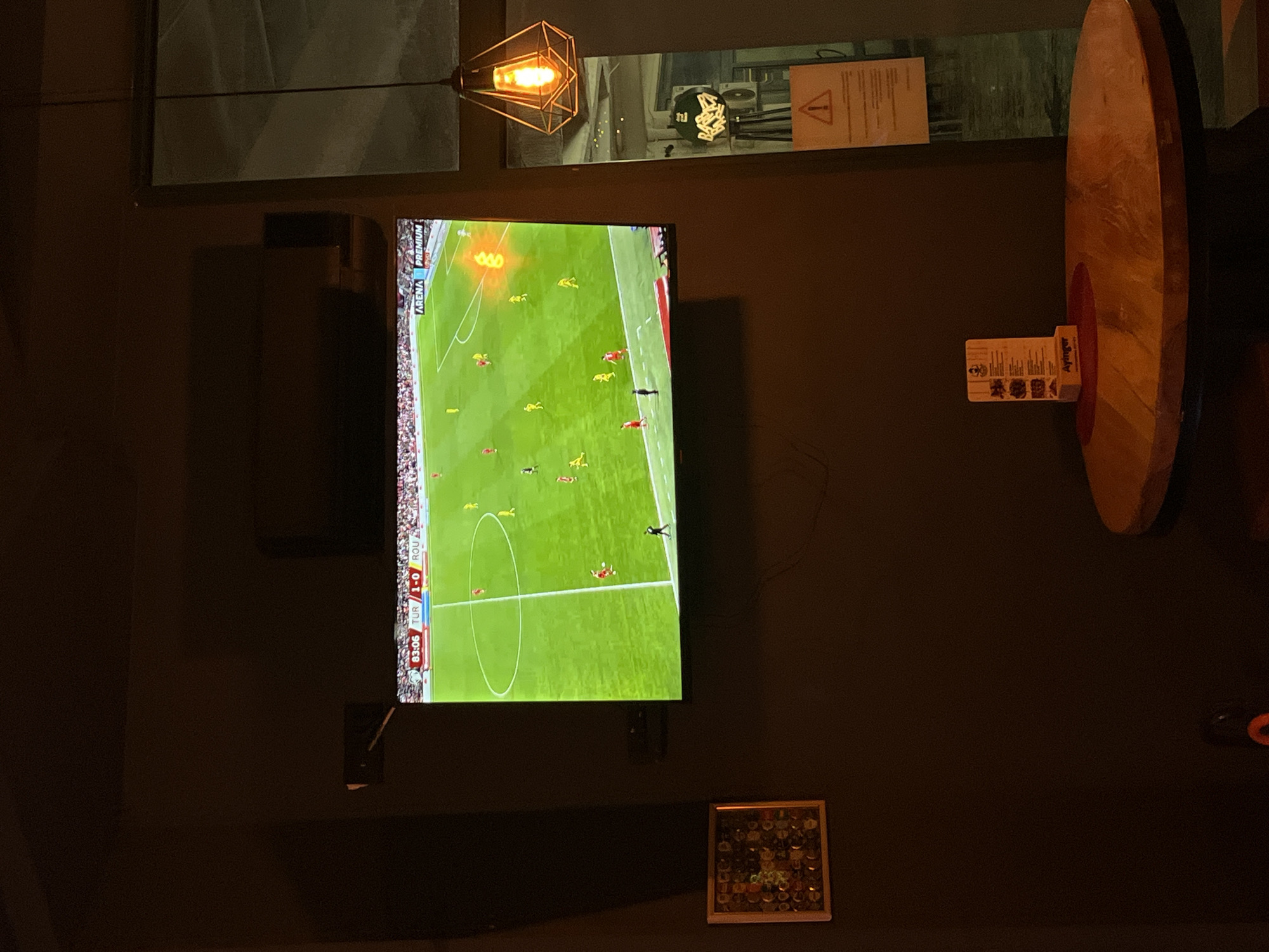 The image displays an interior scene from a establishment identified as Apingər, located in Budva, Montenegro, confirmed by the branding on a menu stand. The setting is dimly lit with dark, possibly brown or charcoal, walls. A large flat-screen television is mounted centrally on the wall, presenting a live soccer match. The on-screen display shows the time as 83:06, with a score of 'TUR 1-0 ROU', and the channel identified as 'ARENA PREMIUM'. Multiple players in red and yellow jerseys are visible on the green soccer field, with a stadium crowd discernible in the background.
To the left and below the television, a square, framed artwork composed of numerous small, circular, multi-colored items, consistent with bottle caps, is affixed to the wall. To the right, a round wooden table with a dark, robust base is positioned. It holds a circular red placemat, upon which a vertical menu stand with the "Apingər" logo is placed. Hanging to the right of the table, a geometric pendant light with a dark metal frame encases an exposed, glowing Edison-style filament bulb, casting a warm light. A tall, rectangular mirror partially reflects elements of the interior, including a yellow warning sign featuring an exclamation mark, and what appears to be a black speaker or equipment mounted on a tripod stand beyond the table. No individuals are directly visible within the immediate foreground of the depicted space.