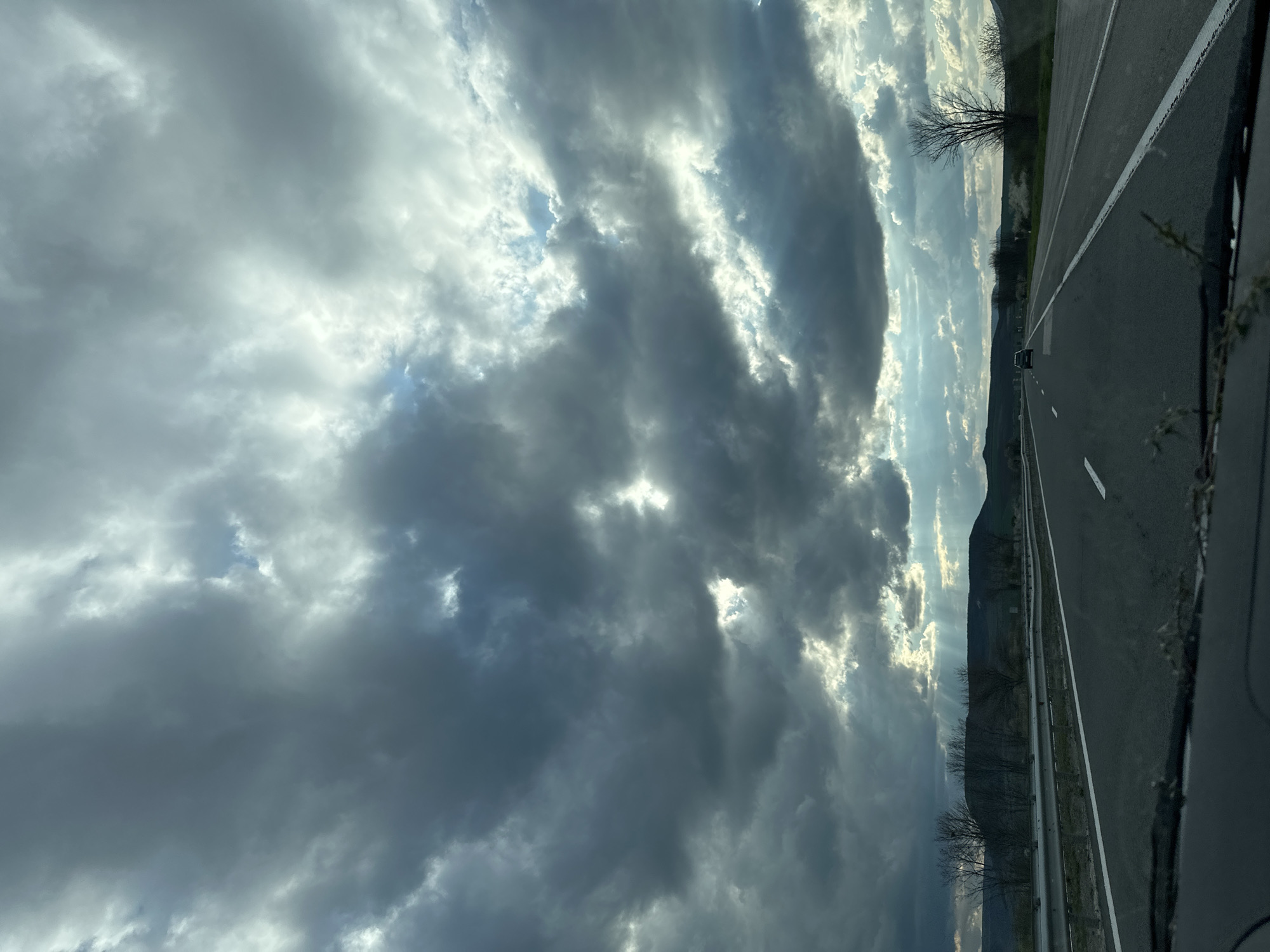 The image depicts a road scene in Zlatina, Bulgaria. A two-lane asphalt road with a white line dividing the lanes is visible. The road is adjacent to a grassy area and a mountain range can be seen in the background. The sky is filled with heavy, dark clouds, with some sunlight breaking through. A car is visible on the road ahead. The photograph appears to have been taken from inside a moving vehicle.