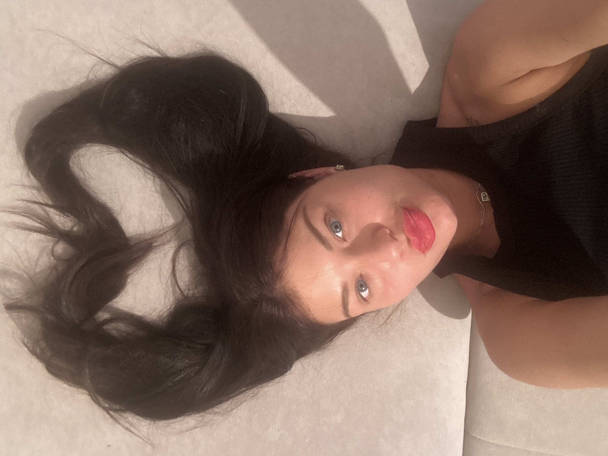 The image captures a young woman lying on a light-colored, soft surface, likely a bed or sofa, in an indoor setting. Her head is turned towards the camera, and she looks directly upwards with striking blue eyes. Her long, dark hair is spread out behind her head, flowing across the light surface. She has dark, defined eyebrows and wears bright red or pink lipstick on her slightly parted lips.
She is dressed in a black ribbed top. Visible accessories include a delicate silver chain around her neck with a small, square pendant, possibly an initial 'B'. A small, shiny earring is also visible in her right ear. Strong, directional lighting from the left casts distinct shadows of her head and hair onto the surface, highlighting the textures and contours. The woman's expression is calm and direct. No text is visible within the image. The contextual location is Moscow, Russia.