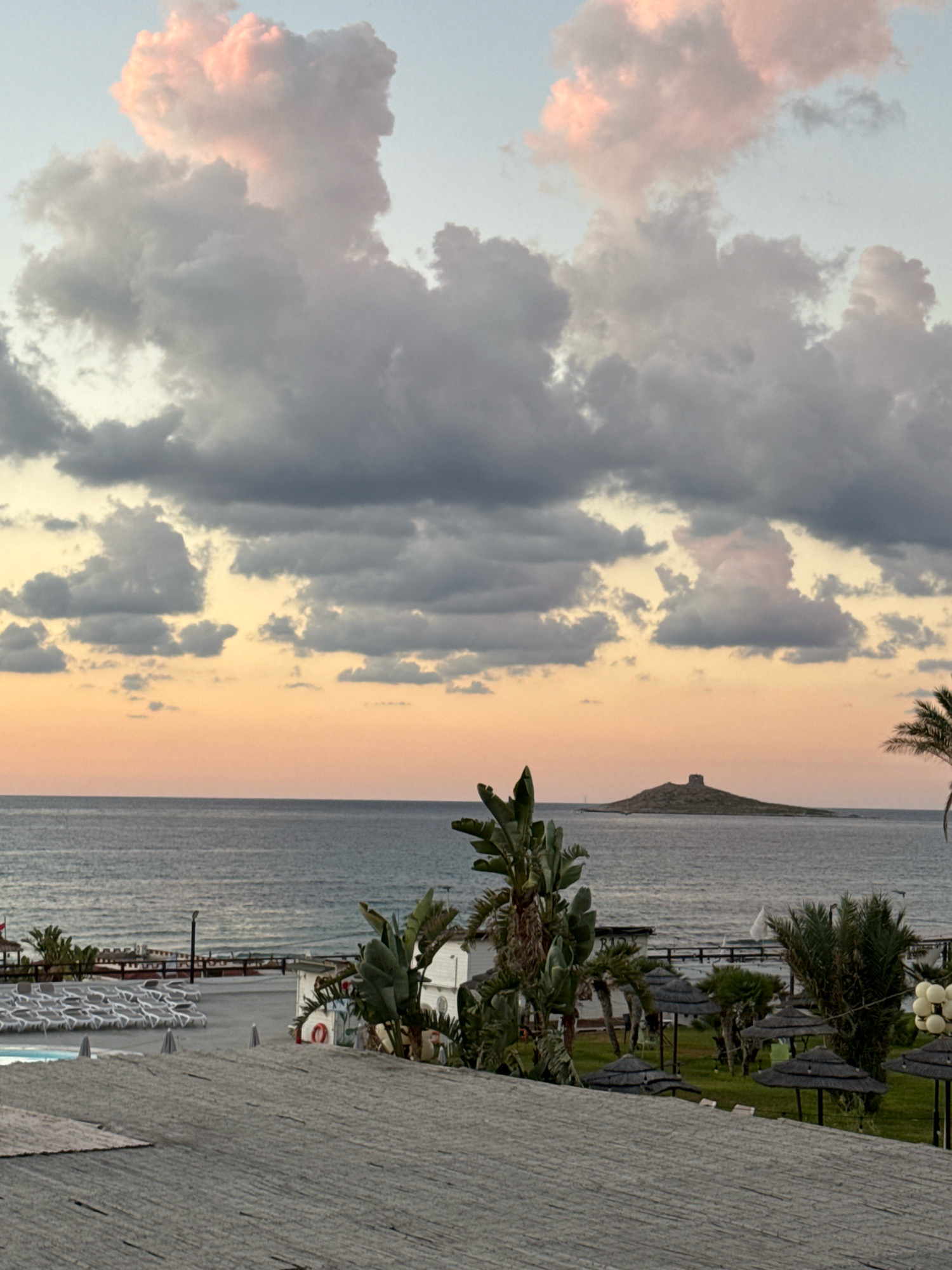 The image depicts a coastal scene in Capaci, Italy, likely during sunset or sunrise. The sky is filled with dramatic clouds, some tinged with pink and orange hues from the low sun, contrasting with darker, gray clouds. Below the sky, a vast expanse of the sea stretches to the horizon.
In the mid-ground, a small, rocky island with a structure resembling a tower or fortification is visible in the distance. Closer to the shore, there are signs of a resort or beach area. Several rows of white lounge chairs are arranged around a swimming pool. There are also thatched umbrellas providing shade over grassy areas. Palm trees and other tropical foliage are scattered throughout the resort. A white building with a red life preserver ring visible on its exterior is also present. The foreground is dominated by a textured, grey surface, possibly a roof or walkway. The overall atmosphere is serene, with the natural beauty of the coast and sky complemented by the resort amenities.