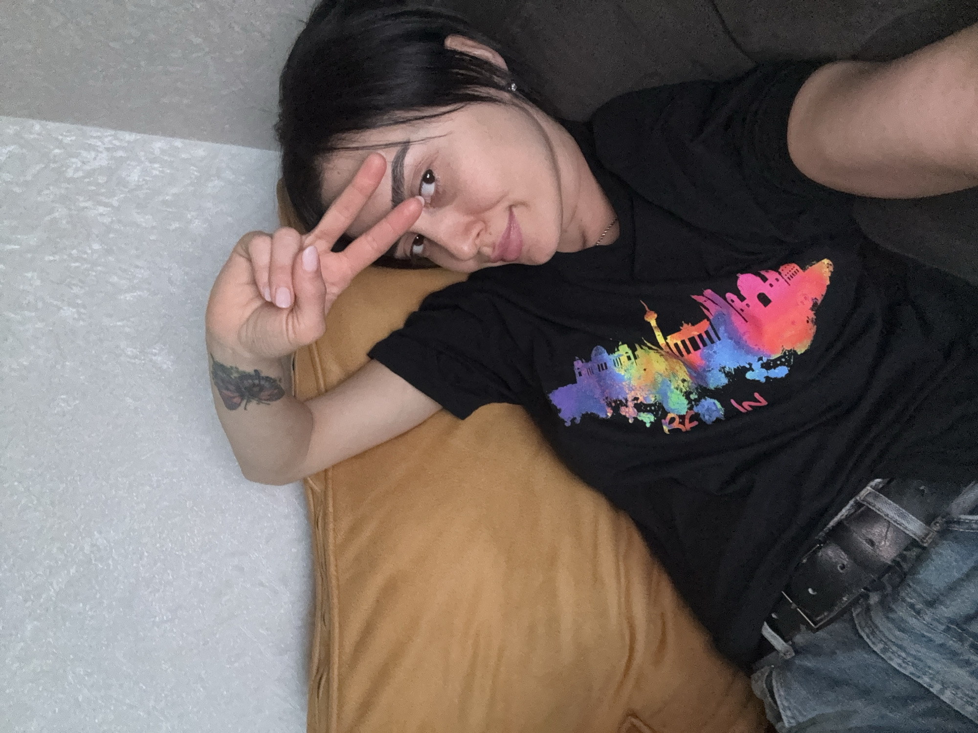 A woman with dark hair is lying on a mustard-colored pillow and a dark couch, making a peace sign with her right hand. She is wearing a black t-shirt with a colorful illustration of Berlin on it and blue jeans with a black belt. There is a butterfly tattoo on her right forearm. The location is unknown and cannot be determined from the image.