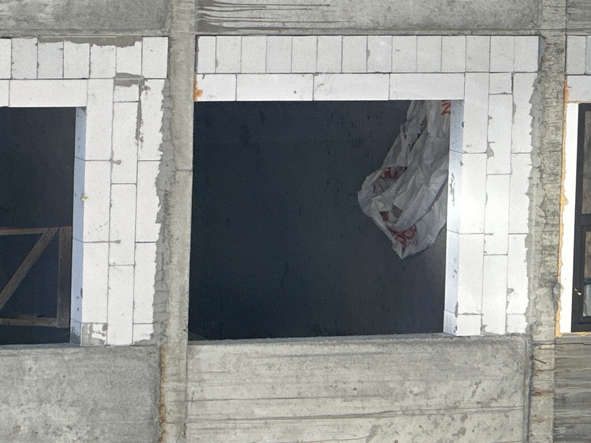 The image shows a close-up view of a partially constructed building with exposed concrete and white aerated concrete blocks. The scene appears to be an outdoor construction site. In the foreground, there are two window openings. The lower window is dark and appears to have some white plastic sheeting with red markings inside. The upper window is also dark and has a wooden frame structure within it. The concrete surfaces show signs of weathering and some staining, possibly from water runoff. The aerated concrete blocks are laid in courses, with some visible mortar between them. The overall impression is of a building in progress, likely in an urban setting given the scale of the structure. The image does not provide specific visual cues to pinpoint its location within the city of Kyiv, Ukraine.