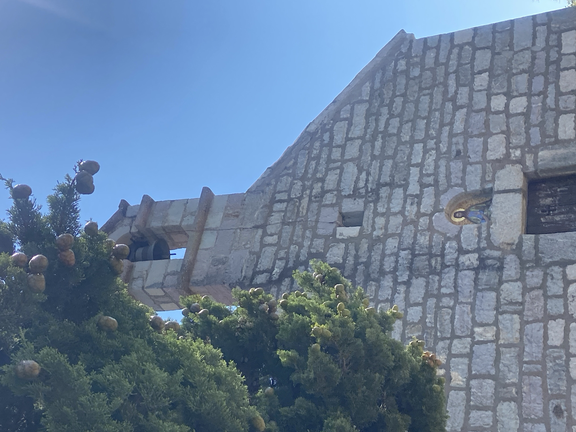 In the village of Blizikuće, Montenegro, a stone building with a bell tower stands against a clear blue sky. The building's exterior is constructed from rough-hewn stone blocks, and a small, arched niche above the entrance contains a mosaic depiction of a religious figure, likely Jesus Christ, with a halo. A dark, weathered wooden door is set into the entrance. The building is partially obscured on the left by a lush green coniferous tree bearing numerous cones. The bell tower, also made of stone, has an open belfry where a dark bell is visible. The overall impression is one of rustic simplicity and a connection to nature.