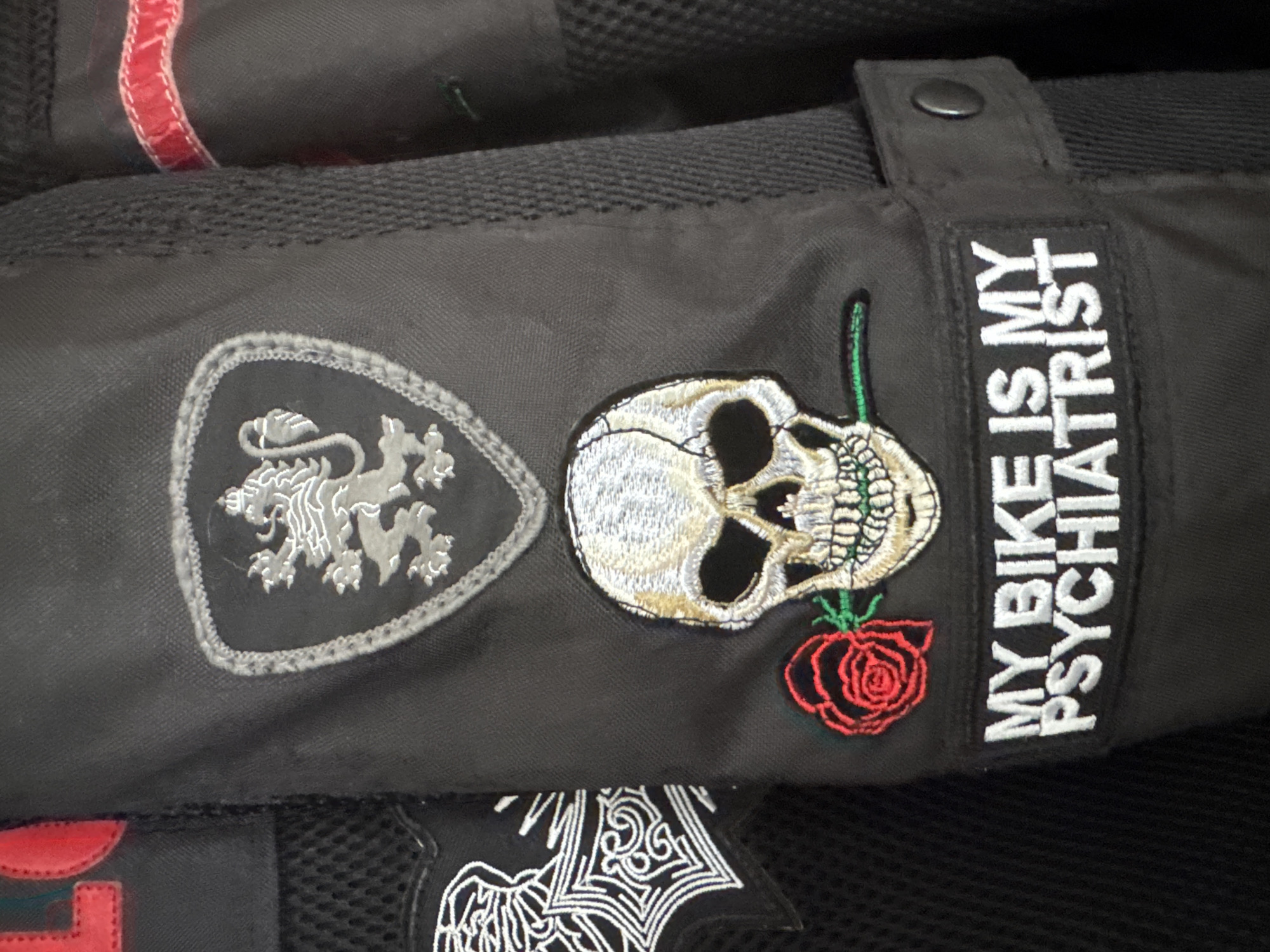 The image shows a close-up of a black garment, possibly a jacket or bag, adorned with several patches. One patch features a grey heraldic lion within a shield-like shape. Adjacent to it is a skull embroidered with white and gold thread, accompanied by a red rose with a green stem. Next to the skull is a rectangular patch with the white text "MY BIKE IS MY PSYCHIATRIST" stitched onto it. Below these patches is a glimpse of another patch with white line work. The overall setting is indeterminate. There is no information to indicate that the location of the image is in Plovdiv, Bulgaria.