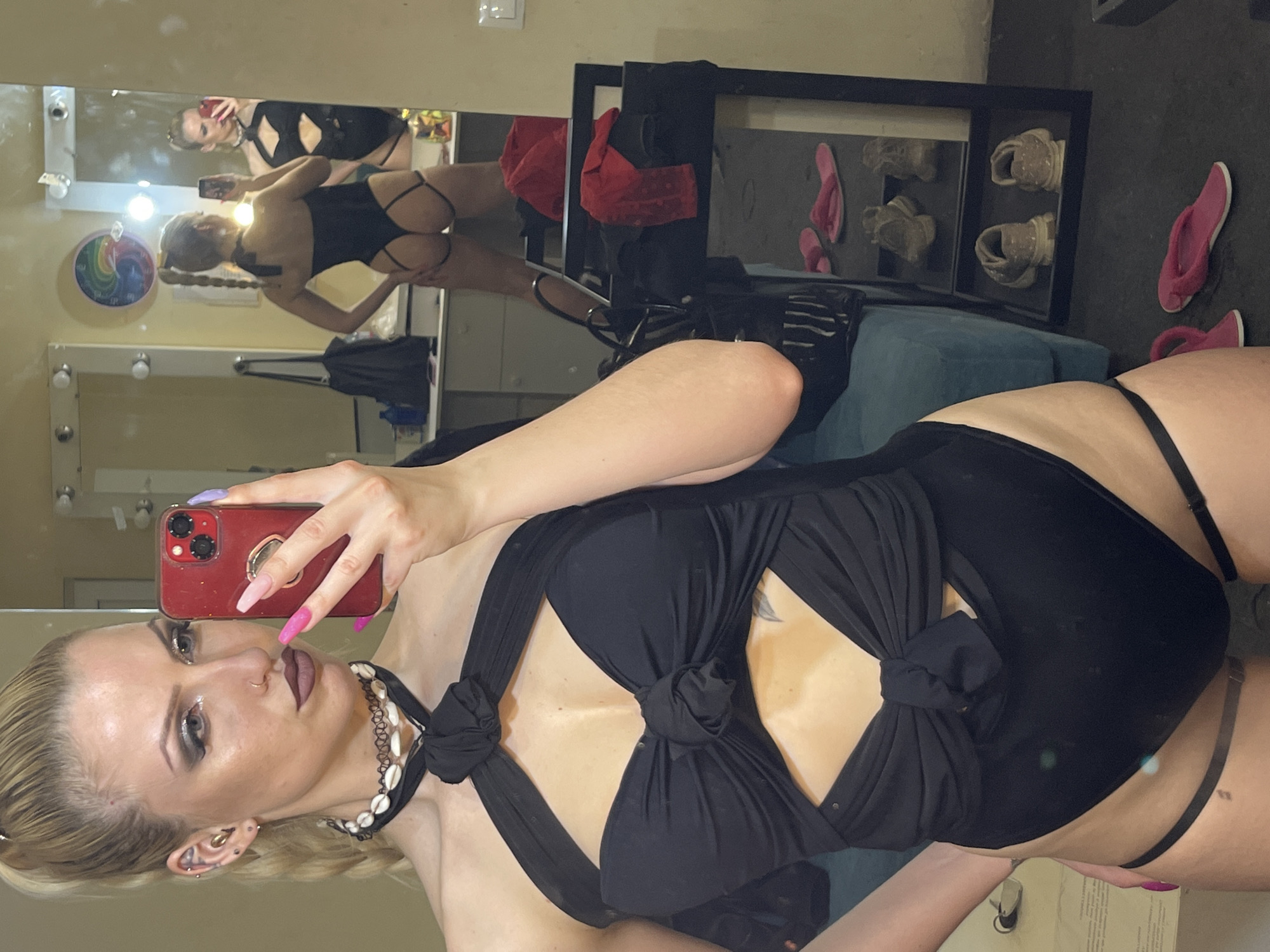 A woman with blonde hair is taking a selfie in a room with a large mirror. She is wearing a black outfit with a tied front and straps around her waist and legs. In the mirror's reflection, another image of the woman wearing a similar outfit can be seen. The room appears to be a dressing or makeup room, equipped with vanity mirrors and lighting. A rack with shoes and bags is visible in the background. The location within the city of Plovdiv, Bulgaria cannot be determined from the information in the image.