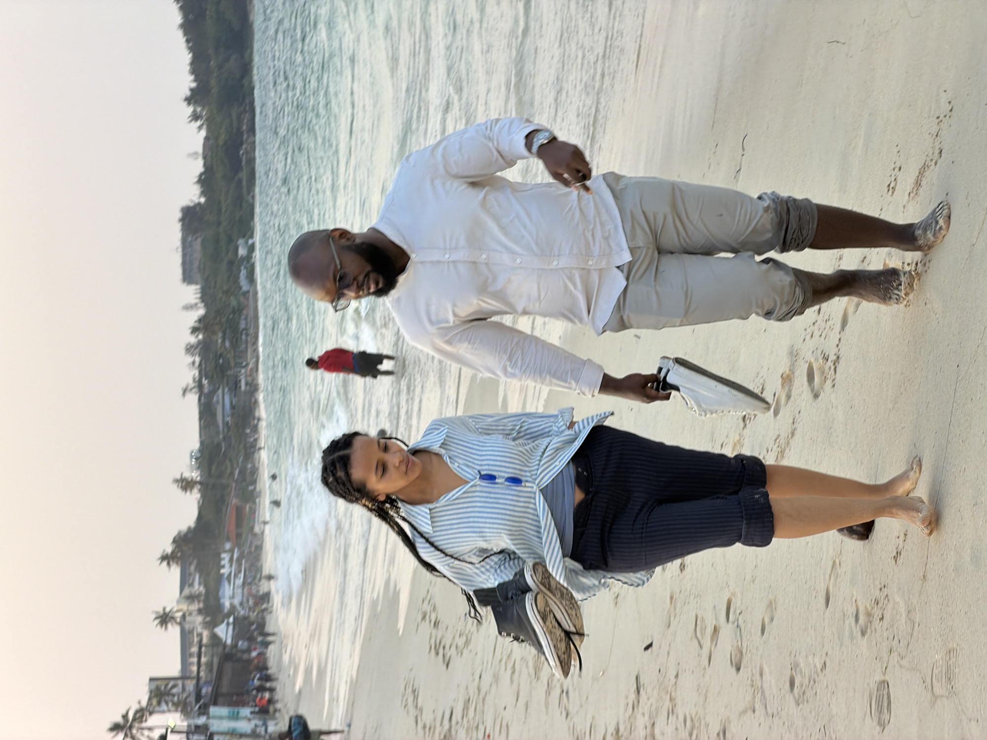 Two individuals, a man and a woman, walk barefoot on a sandy beach along the coast of the Indian Ocean, likely near the city of Dar es Salaam, Tanzania. The woman, positioned in the foreground on the left, is carrying a pair of black high-top sneakers in her left hand. She is wearing a light blue and white striped button-down shirt, navy blue capri pants, and has her hair styled in braids. The man, to her right, is wearing a light-colored collared shirt and light-colored capri pants. He is carrying a white sneaker in his right hand. The water is visible behind them, with gentle waves lapping at the shore. In the background, other people are visible on the beach, as well as some buildings and palm trees lining the coast. The sky is clear and appears to be either early morning or late afternoon, with soft, diffused light. The scene depicts a leisurely stroll along a tropical beach.