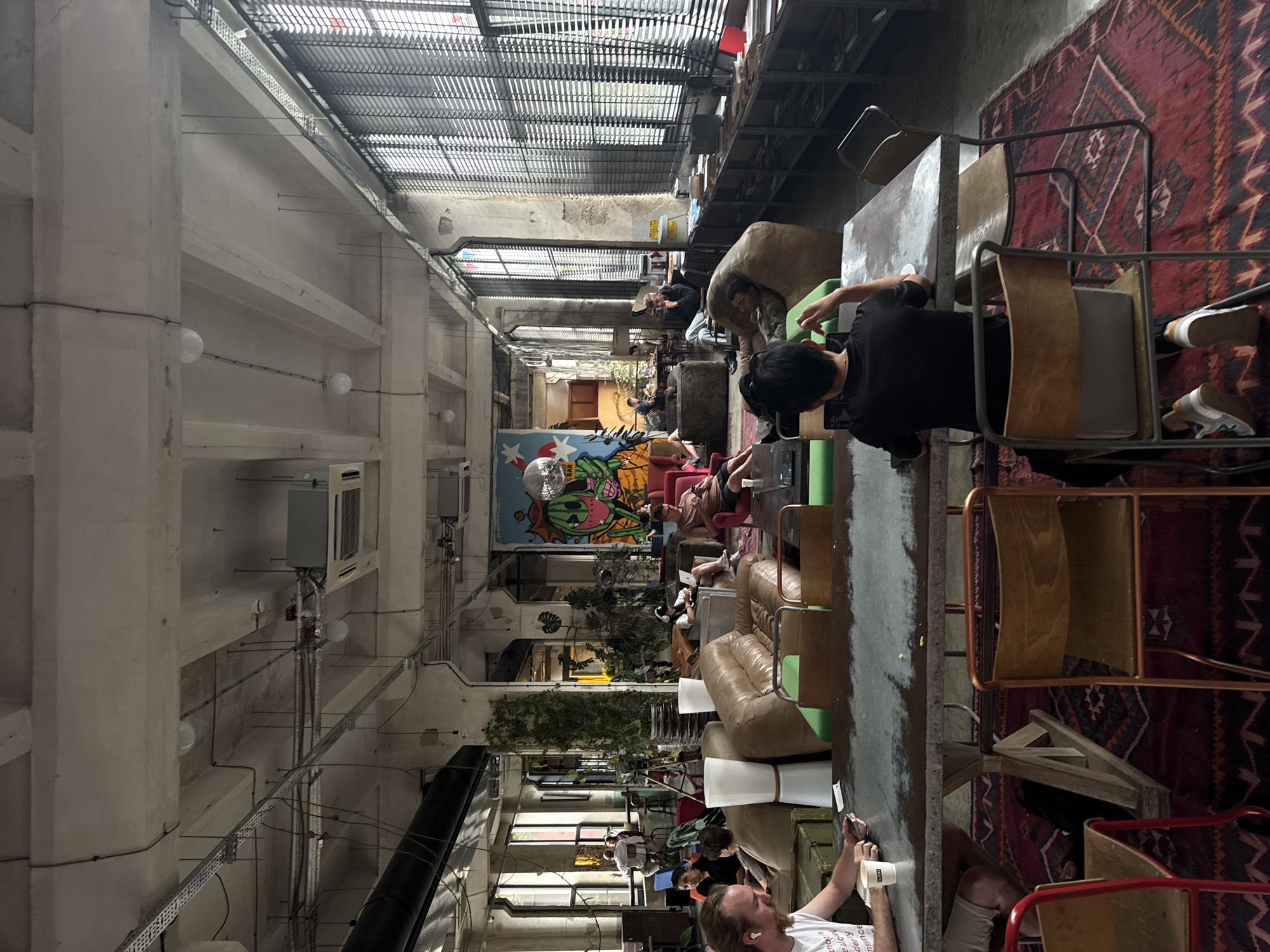 This image depicts the interior of a spacious, industrial-style co-working space in Tbilisi, Georgia. The room is characterized by its high ceilings with exposed concrete beams and ductwork, large windows with blinds, and a mix of modern and vintage furniture. The environment is brightly lit, with natural light filtering through the windows and overhead lighting.
Several people are present, engaged in various activities typical of a co-working environment. In the foreground, a man with a beard and white t-shirt is seated at a large metal table, holding a phone and a cup. Across from him, a person in a black t-shirt is working on a laptop. Other individuals are scattered throughout the space, some lounging on couches, others working at tables, and some conversing.
The furniture includes large, plush leather sofas, beanbag chairs, wooden chairs, and metal-framed tables. The decor is eclectic, featuring large potted plants, a colorful mural on the wall, and a disco ball. Shelving units filled with books and other items line one wall. The overall impression is one of a dynamic and collaborative workspace, likely a popular spot for freelancers and remote workers in Tbilisi.