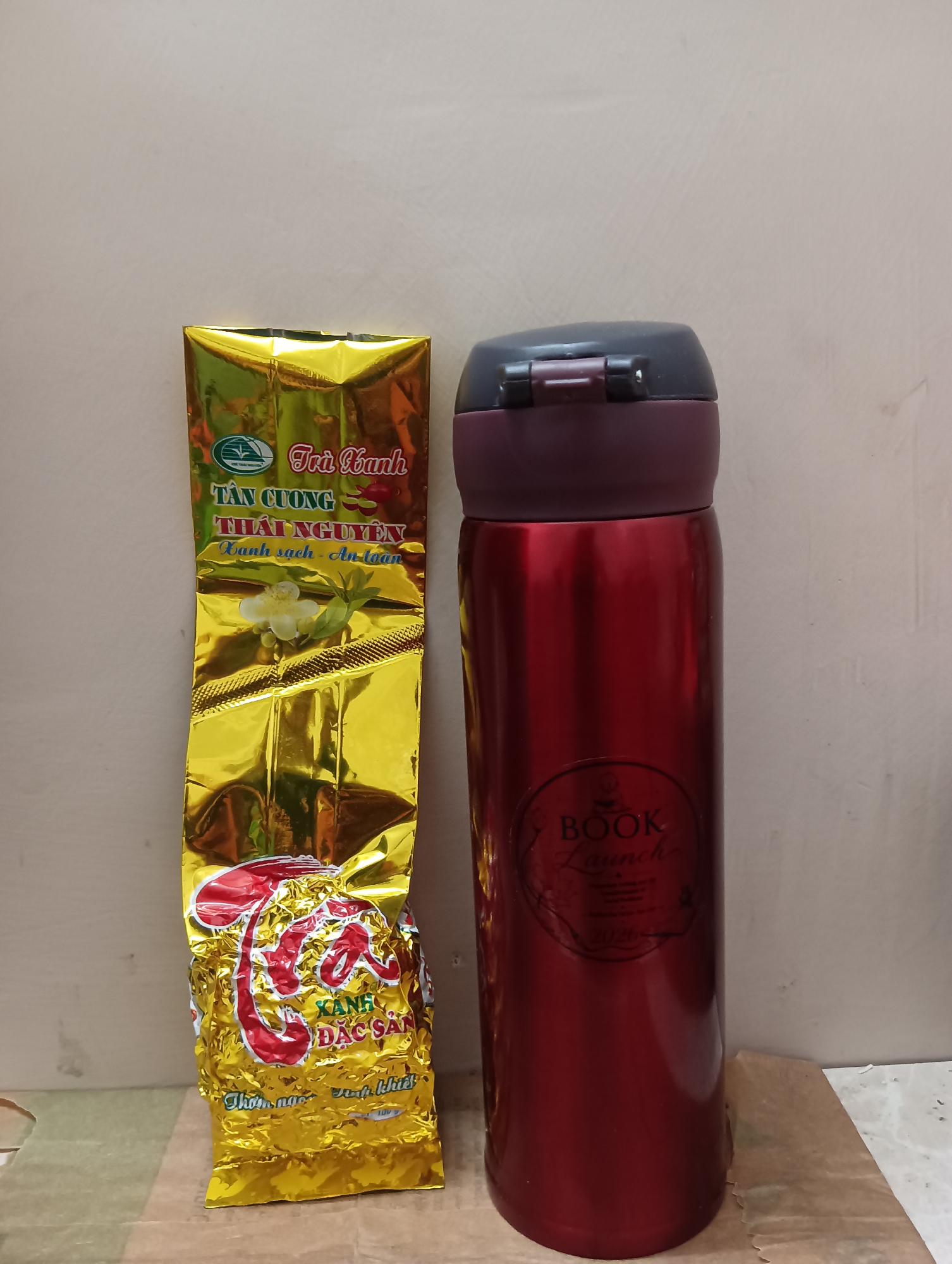 In the foreground of the image, a tall, slender, metallic red thermos bottle stands upright on a light brown surface. It has a dark gray lid with a hinged opening mechanism. To the left of the thermos, a long, crinkled, golden foil package of tea is leaning against it. The tea package has red and green text and graphics, including an image of a white flower with green leaves. The background is a textured, light-colored wall. The scene is illuminated by artificial light, casting shadows on the wall. The overall setting appears to be indoors, on a table or shelf. There is no indication of Manali, India, or any outdoor elements.