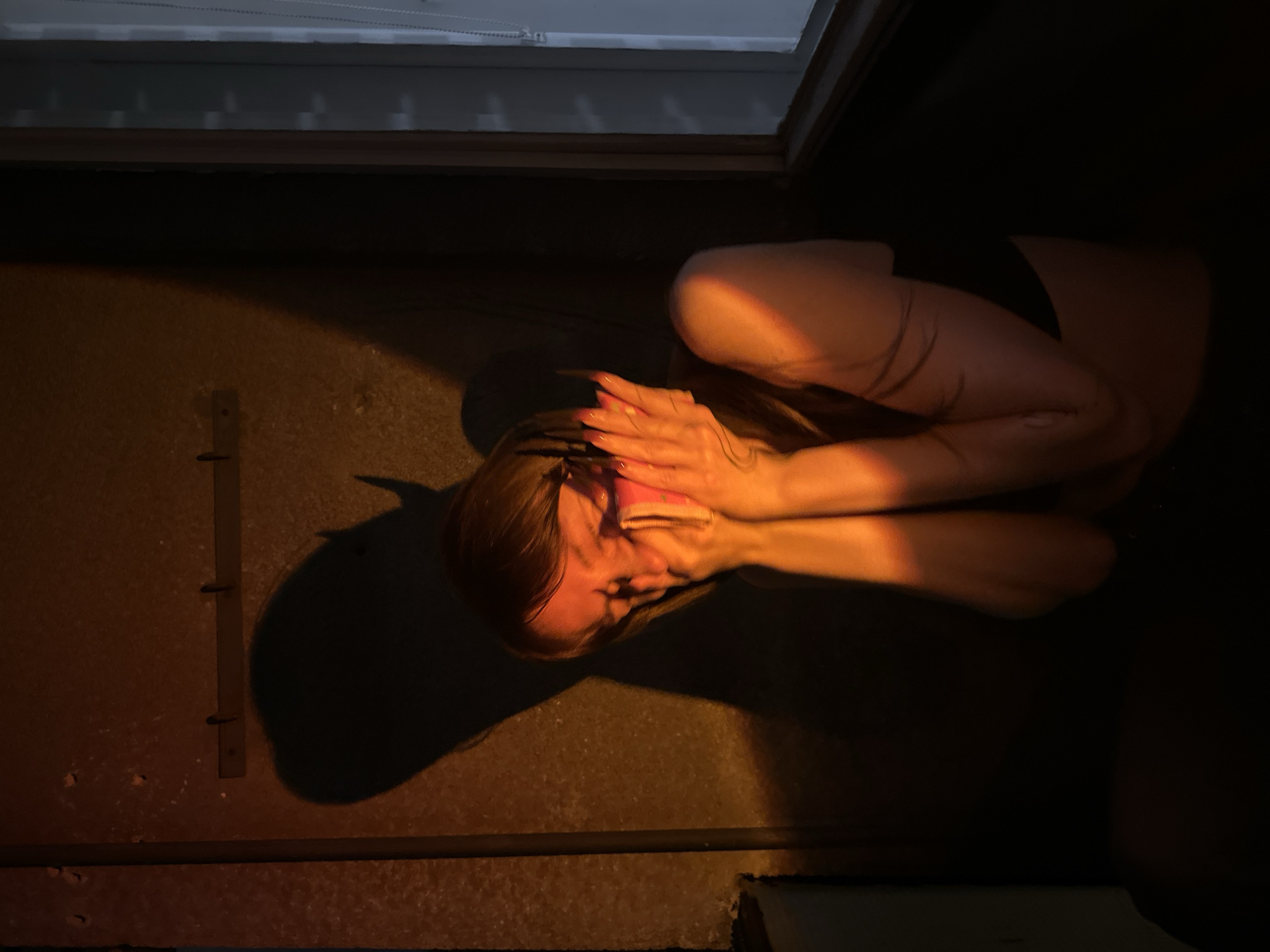 A person is crouched on a concrete floor, their face obscured by their hands. The area is dimly lit with a golden light source casting a shadow of the person's head onto the floor. A metal grate and a window are also visible. The location appears to be inside a building. There is no definitive evidence to suggest this image was taken in the city of Vracar Urban Municipality, Serbia.