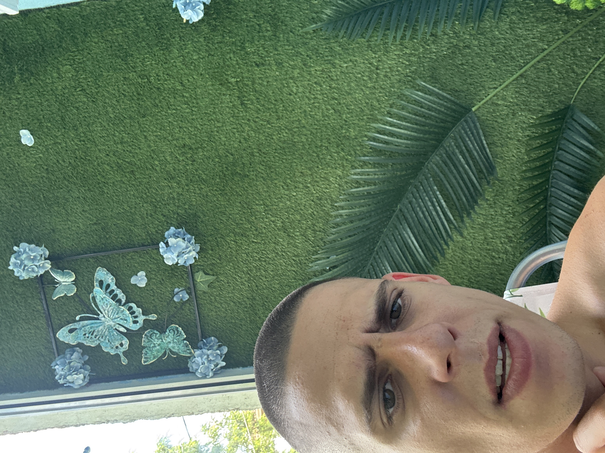 A man with a buzz cut and brown eyes is taking a selfie. He is looking at the camera with a furrowed brow and his mouth is slightly open, revealing his teeth. He is wearing a white tank top. The background is a wall covered in artificial green grass. There are several light blue hydrangea flowers and butterfly decorations on the wall, as well as some artificial palm leaves. The man's left arm and shoulder are visible. The scene appears to be outdoors, possibly in a garden or patio area. The location is not specified.