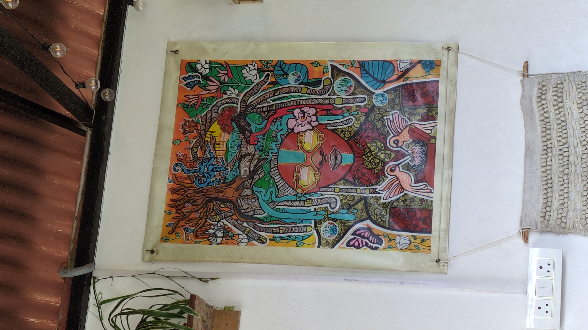 A framed artwork depicting a person with elaborate dreadlocks, adorned with yellow sunglasses and facial markings, hangs on a white wall in Haini, India. The artwork features a vibrant, psychedelic style with elements like birds, butterflies, and flora. Below the artwork, a woven textile hangs from a wooden dowel. To the left of the textile and artwork, a white electrical socket with three circular receptacles and two switches is mounted on the wall. Above, a corrugated brown roof is supported by black beams, with string lights hanging from the beams. A potted plant with long green leaves is visible on a wooden shelf to the left.
