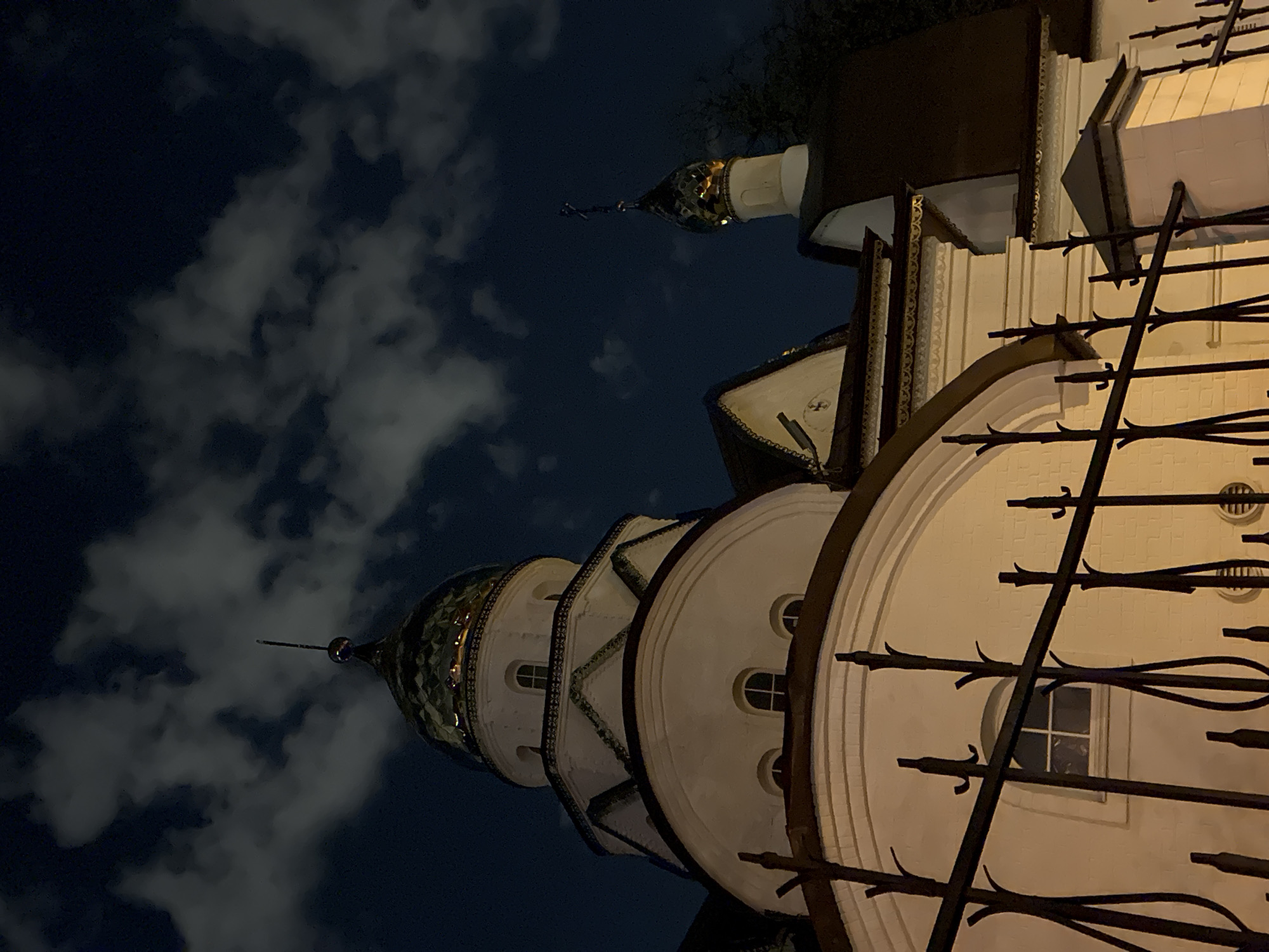 The image shows the Novodevichy Convent in Moscow, Russia, at night. The convent is a historic fortress and monastic complex. The architectural elements of the convent are visible, including the tiered towers with domes. In the foreground are wrought iron railings. The sky is dark with scattered clouds.
