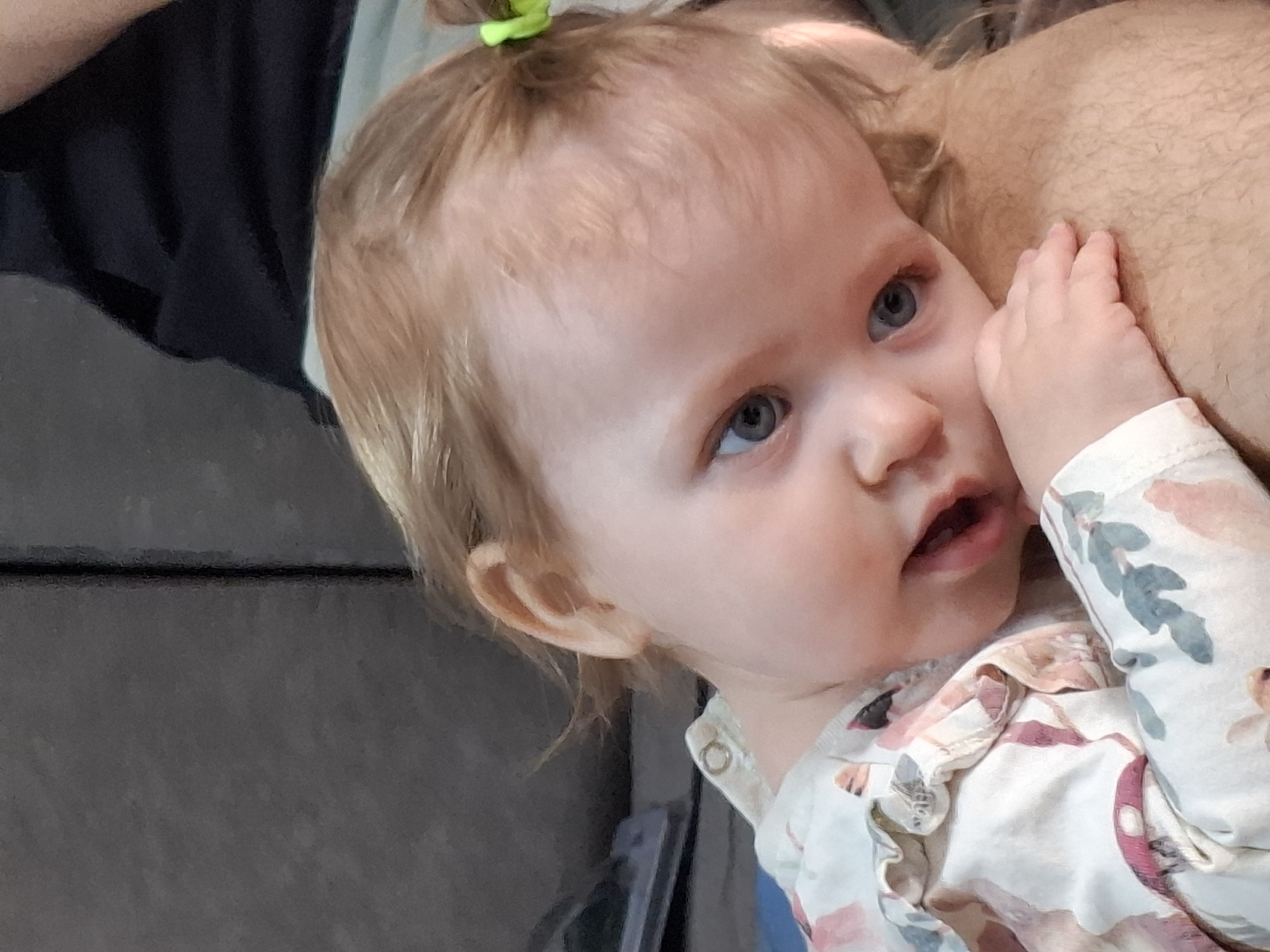 A close-up shot depicts a baby with fair skin and blue eyes. A small green hair tie is visible atop the baby's light-colored hair. The baby wears a floral-patterned shirt and is nestled against a light brown-skinned adult whose arm is around the child. The baby's hand rests against the adult's skin. Part of a dark gray sofa is visible in the background. The location is unknown and there is nothing present to indicate the location of Chaikovsky, Russia.
