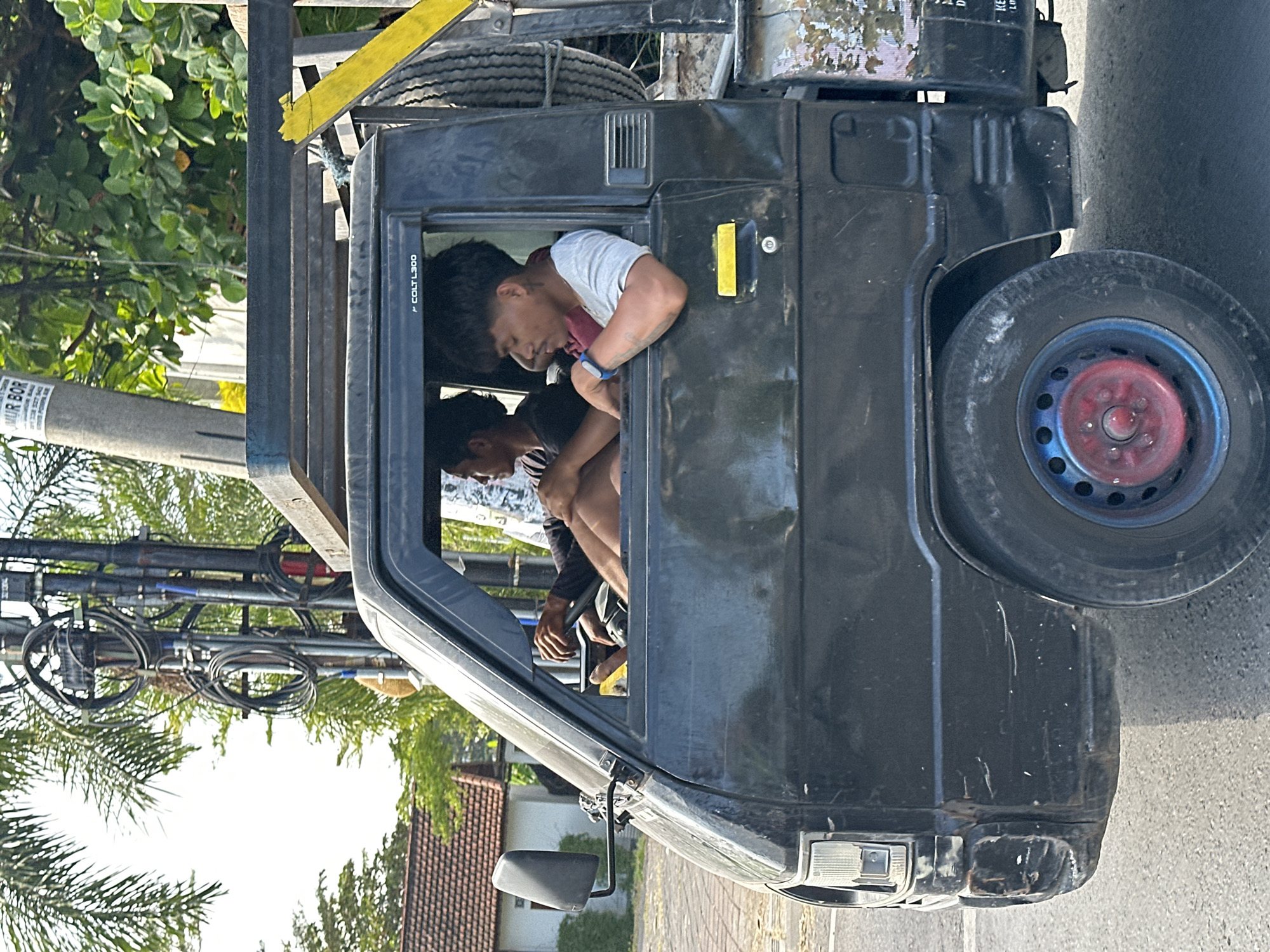 In North Kuta, Indonesia, a black Mitsubishi Colt L300 pickup truck is captured in profile. The truck is in motion, with its front passenger side visible. Two men are inside the cab. The driver, on the left, wears a dark long-sleeved shirt and has his hands on the steering wheel. The passenger, on the right, wears a light-colored t-shirt and rests his arm on the open window frame. His head is turned slightly to the left, and he appears to be looking forward or at the driver. A spare tire is visible mounted on the exterior of the truck's rear bed. The truck's exterior shows signs of wear, with scuffs and scratches on the bodywork. Behind the truck, a utility pole with tangled wires stands tall, and lush green foliage and a tiled-roof building suggest a tropical environment. The road surface is asphalt.