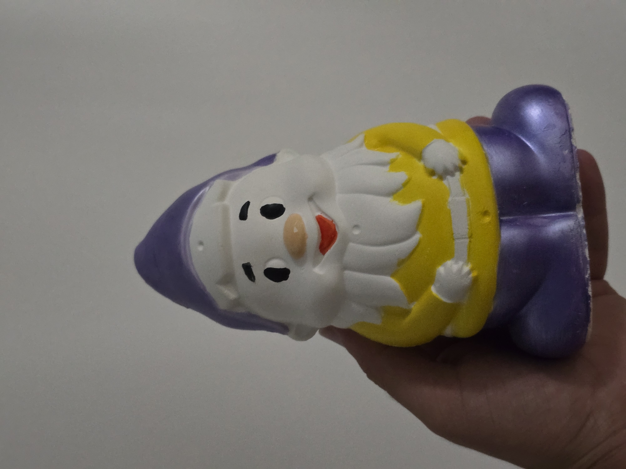 A hand is holding a small, cartoonish gnome figure in front of a plain, light gray background. The gnome is predominantly white with a purple hat and purple pants. It has a yellow tunic with white trim at the collar and a white belt. The gnome's face is white with small black eyes, a peach-colored nose, and a red mouth. Its beard and hands are also white. The figure appears to be made of a soft, possibly squishy material. The background is featureless and evenly lit, suggesting an indoor setting. The image does not contain any information about the city of Qaratau, Kazakhstan.
