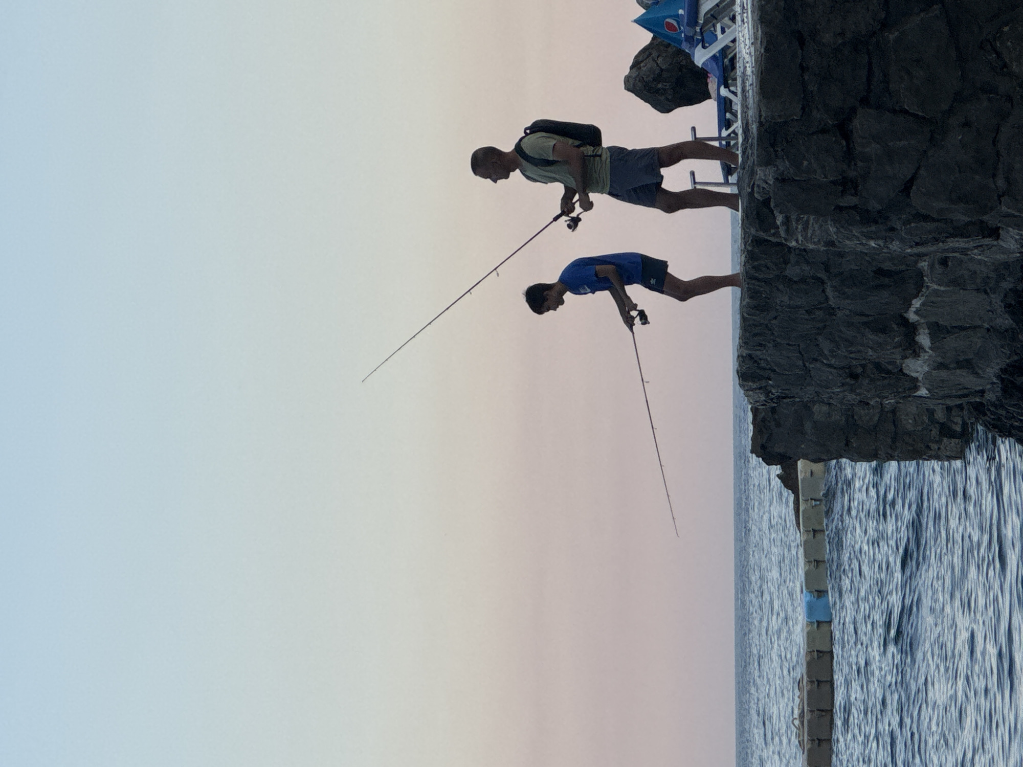 Two individuals, a man and a boy, are silhouetted against a pale pink and blue sky, fishing from a stone pier. The man, wearing a t-shirt and shorts, holds a fishing rod. The boy, in a blue t-shirt and shorts, also fishes. The sea, with gentle waves, extends to the horizon. In the background, a concrete barrier with blue accents is visible in the water, along with lounge chairs and a large rock formation on the pier. The location is Prijevor, Montenegro.