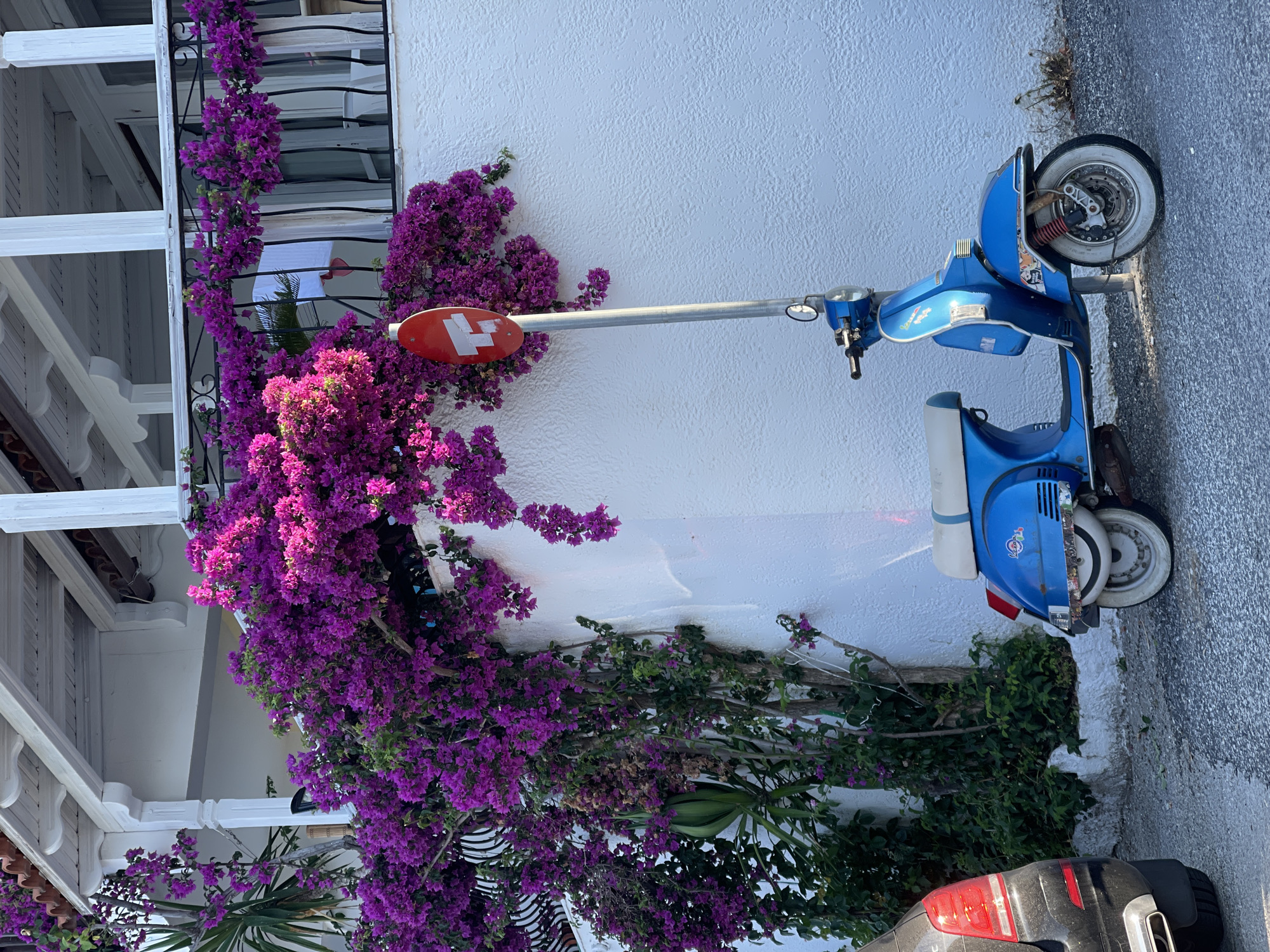 A blue vintage scooter is parked next to a white textured wall. The scooter has a white seat with a blue stripe and white tires. Above the scooter, a red circular sign with white horizontal bars is mounted on a metal pole. Vibrant purple bougainvillea flowers cascade down from an overhang, partially obscuring the wall and the sign. The scooter is positioned on a gravelly surface. In the background, a portion of a black car is visible on the left side of the frame. The scene appears to be outdoors, likely in a street or alleyway. The architecture suggests a Mediterranean setting. The location is the Sithonia Municipal Unit, Greece.