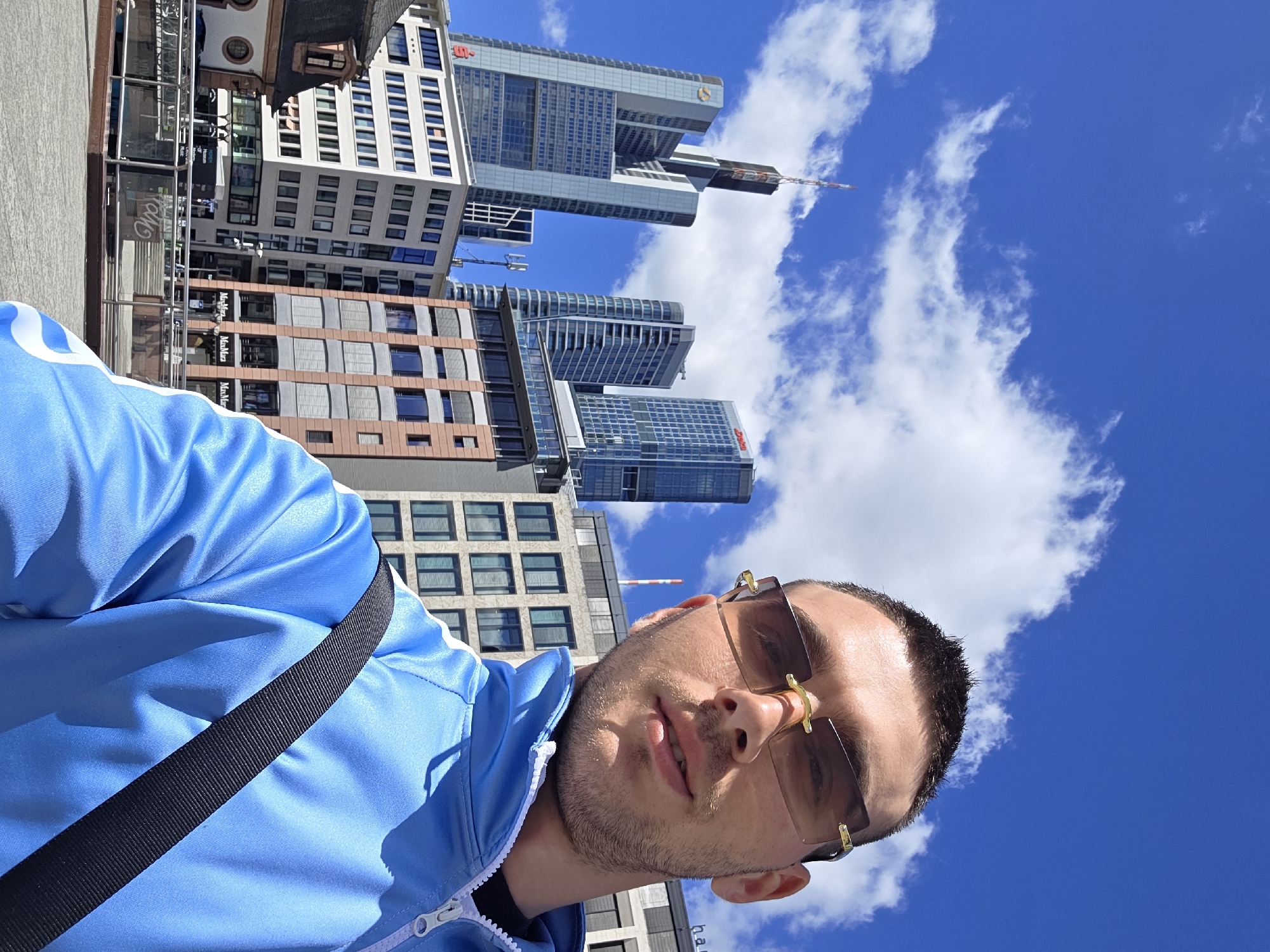 A young man takes a selfie outdoors with a city skyline in the background under a bright blue sky with scattered clouds. The man is wearing a light blue tracksuit with white stripes and gold-rimmed sunglasses. He has short, dark hair and stubble. He is wearing a black strap across his chest. In the background, several modern skyscrapers of varying heights and designs are visible, with some featuring reflective glass facades. One prominent building has a red logo on its upper section. Below the skyscrapers, there are lower buildings, some with patterned facades and windows. A street or plaza area with paved ground is visible in the foreground. The overall scene depicts an urban environment on a sunny day.