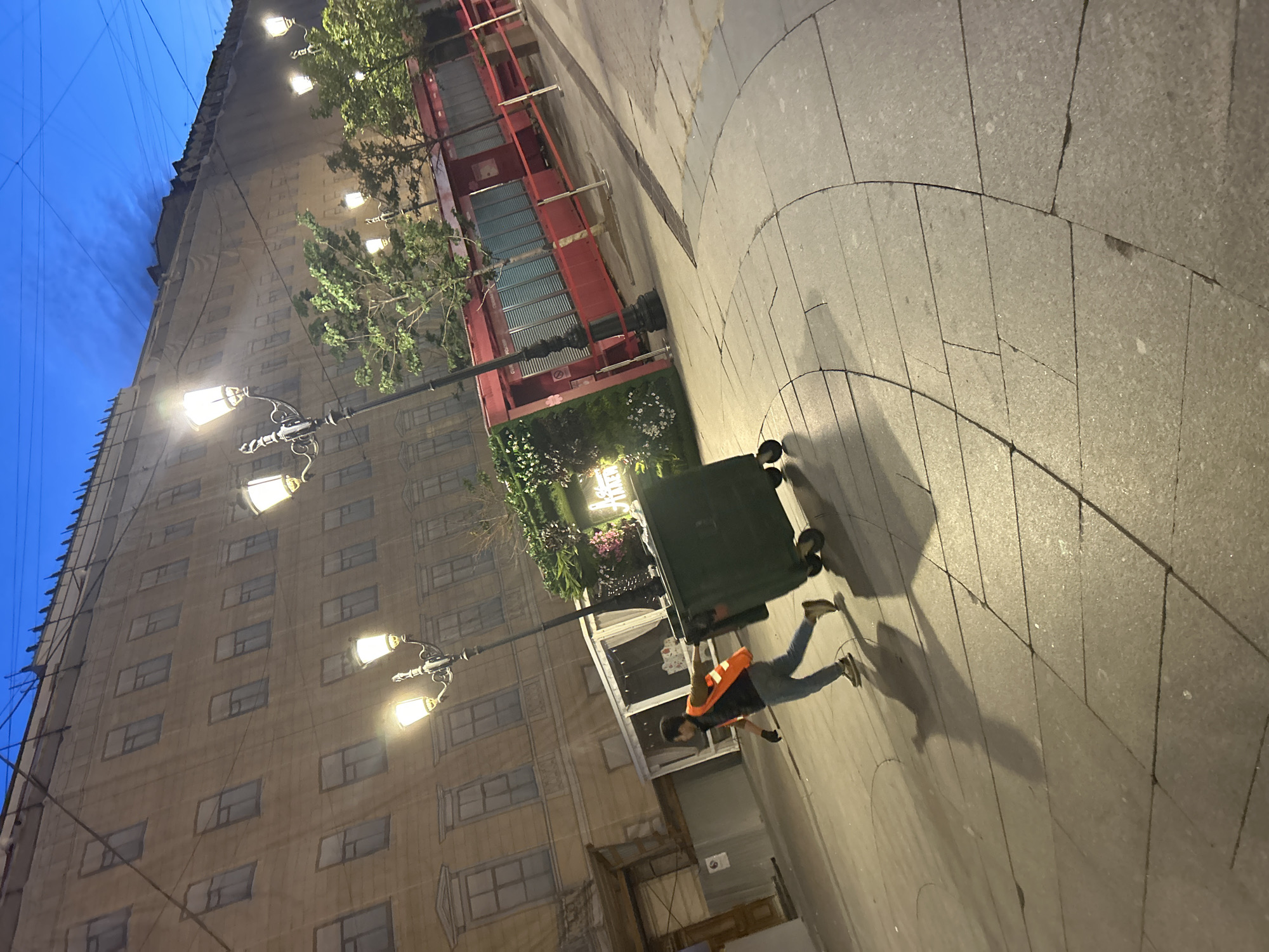 An over-the-shoulder medium shot captures a person in an orange safety vest and blue jeans, pushing a large, dark green trash bin with wheels along a stone-paved sidewalk. The person's face is obscured, and they appear to be in motion, with their body angled as they push the bin. The bin is positioned in the foreground, slightly to the right of the frame, and casts a long shadow. In the background, a building facade is covered with a tarp, creating a flattened, painted appearance of windows and architectural details. Several ornate streetlights with illuminated bulbs are visible, interspersed with the branches of trees. To the right, there are small red kiosks with corrugated metal shutters and red tape barriers. The sky above is a deep twilight blue, with visible power lines crisscrossing the frame. The scene suggests a public urban space, possibly a street or promenade, during the evening or early morning hours.