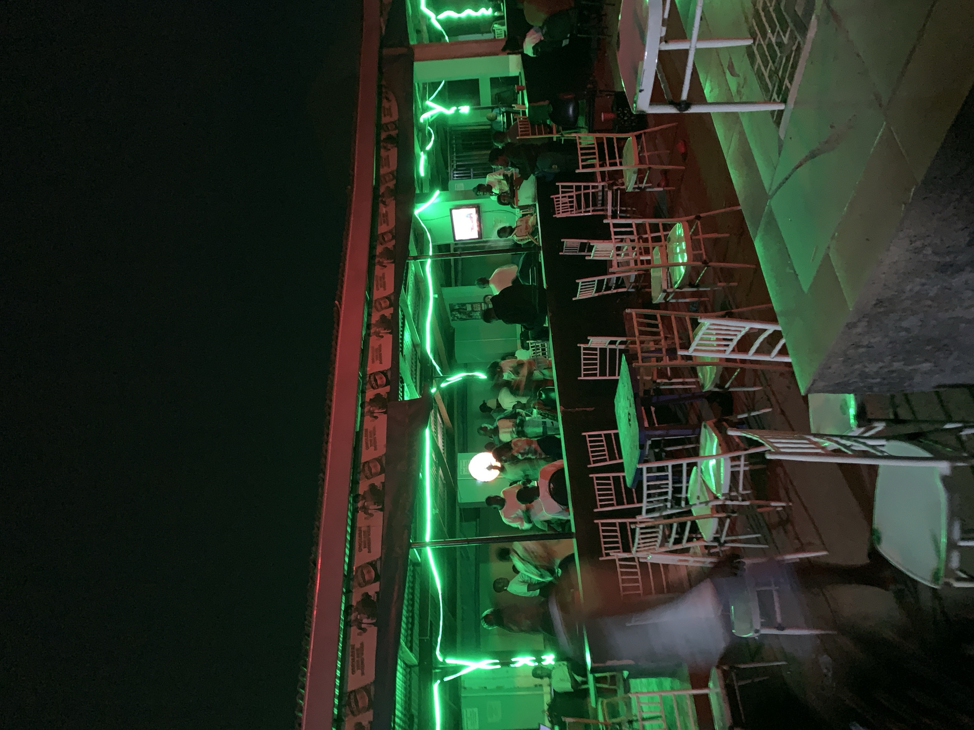 At New Stadium, Nigeria, an outdoor bar scene is captured at night. The location features multiple levels, with patrons seated at tables and chairs. Green neon lighting illuminates the area. A television screen is visible on one of the levels. There are many tables and chairs, most of which are empty and arranged. The bar appears to be open and populated with people. The perspective of the photo is angled. The upper-left portion of the image is dark, while the illuminated bar section occupies the remaining space.