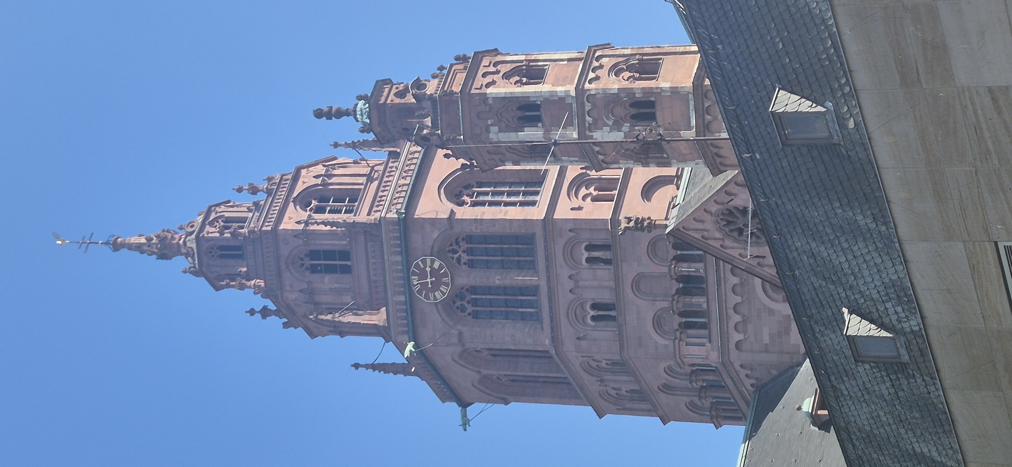 The image shows the tower of the Mainz Cathedral (Mainzer Dom), officially the Cathedral of Saint Martin of Mainz, located in Mainz, Germany. The cathedral is a Romanesque-style building made of red sandstone, and its tower is a prominent feature of the city's skyline. The tower has a clock face visible, and several arched windows. The sky is clear and blue. In the foreground, parts of other buildings with tiled roofs are visible, partially obscuring the base of the cathedral tower. The overall scene depicts an architectural landmark against a clear sky.